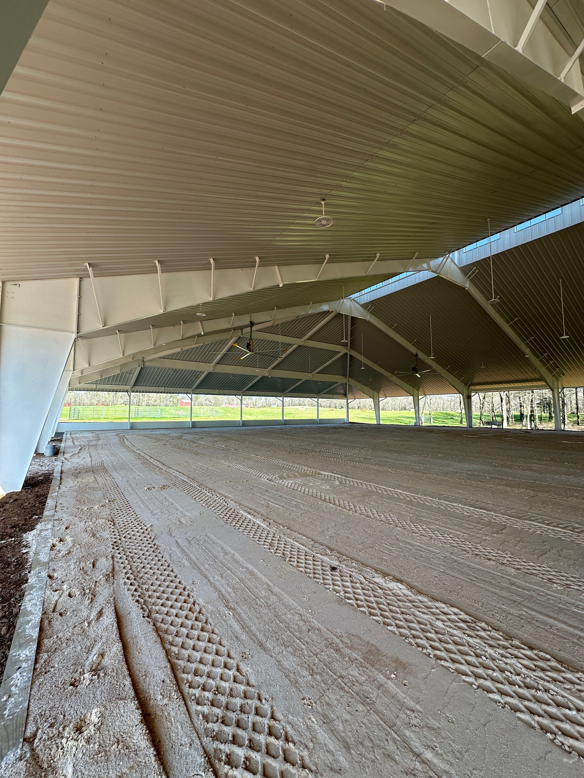 An indoor horse riding arena with a white metal roof, lights, and a sandy track with visible grid-pattern.
