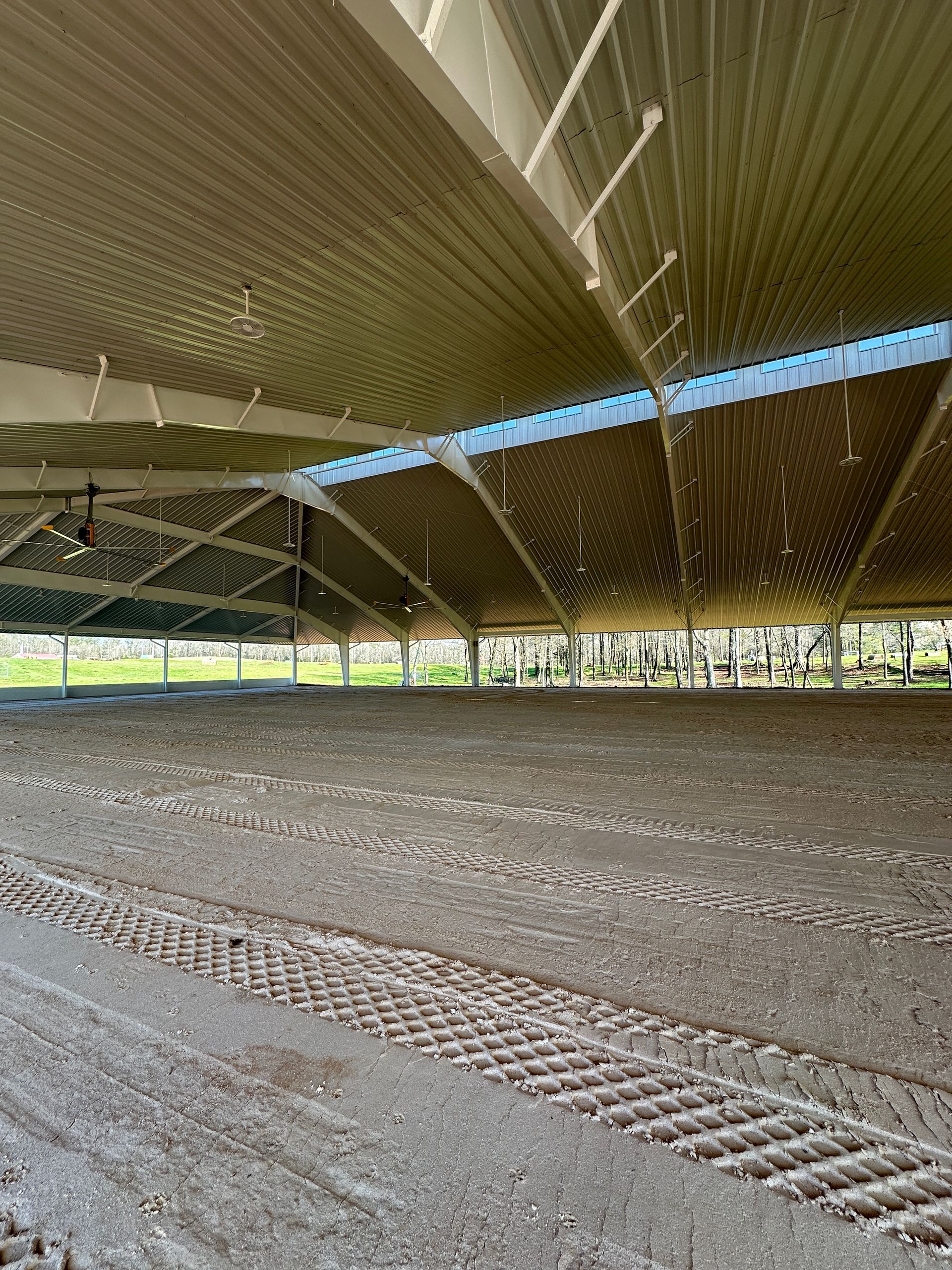 An open-sided equestrian riding arena with a large, high metal roof and a freshly groomed dirt floor with tire tracks.