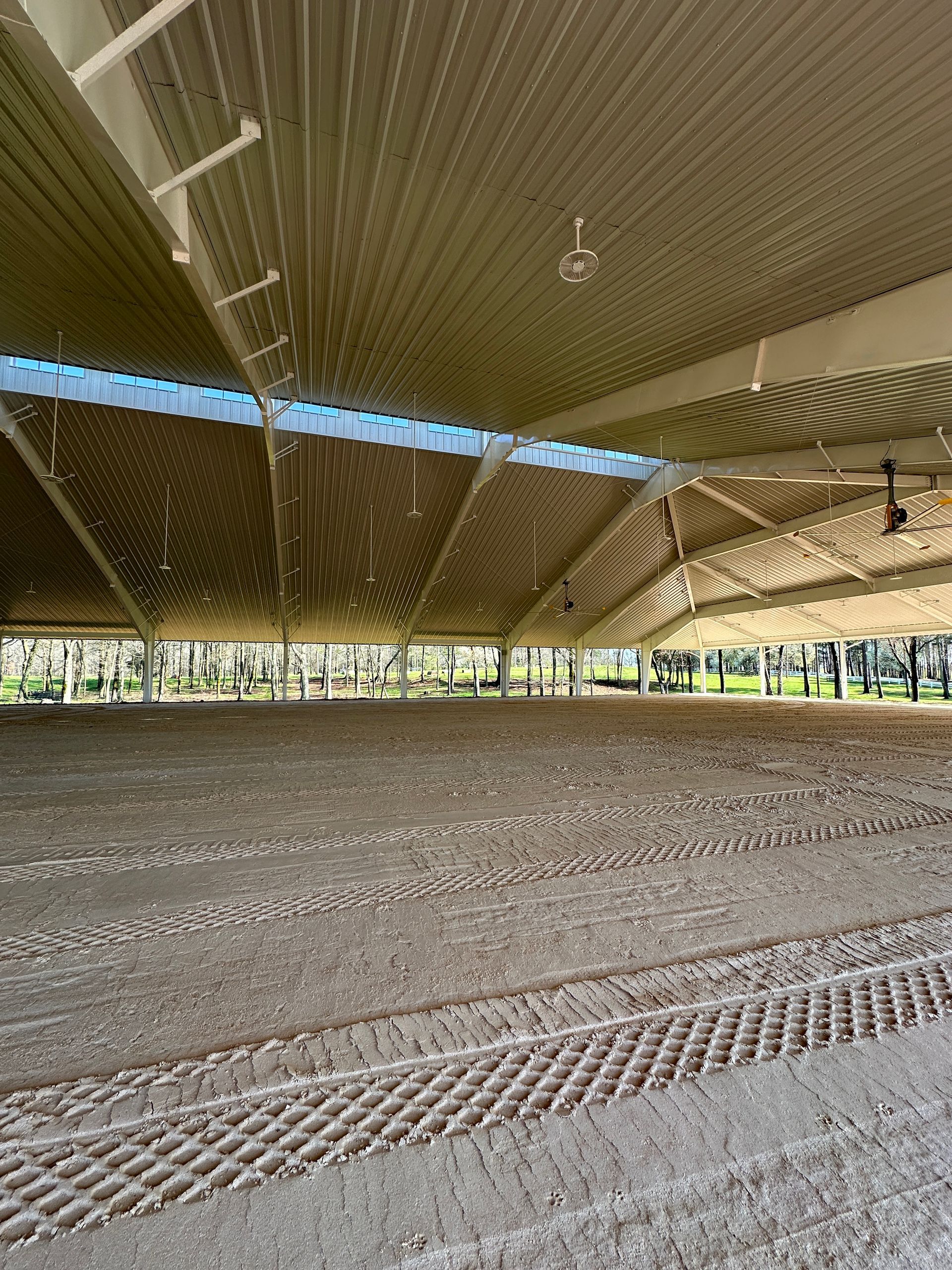 A spacious, covered outdoor equestrian arena with a metal roof, open sides, and a groomed dirt ground surface.