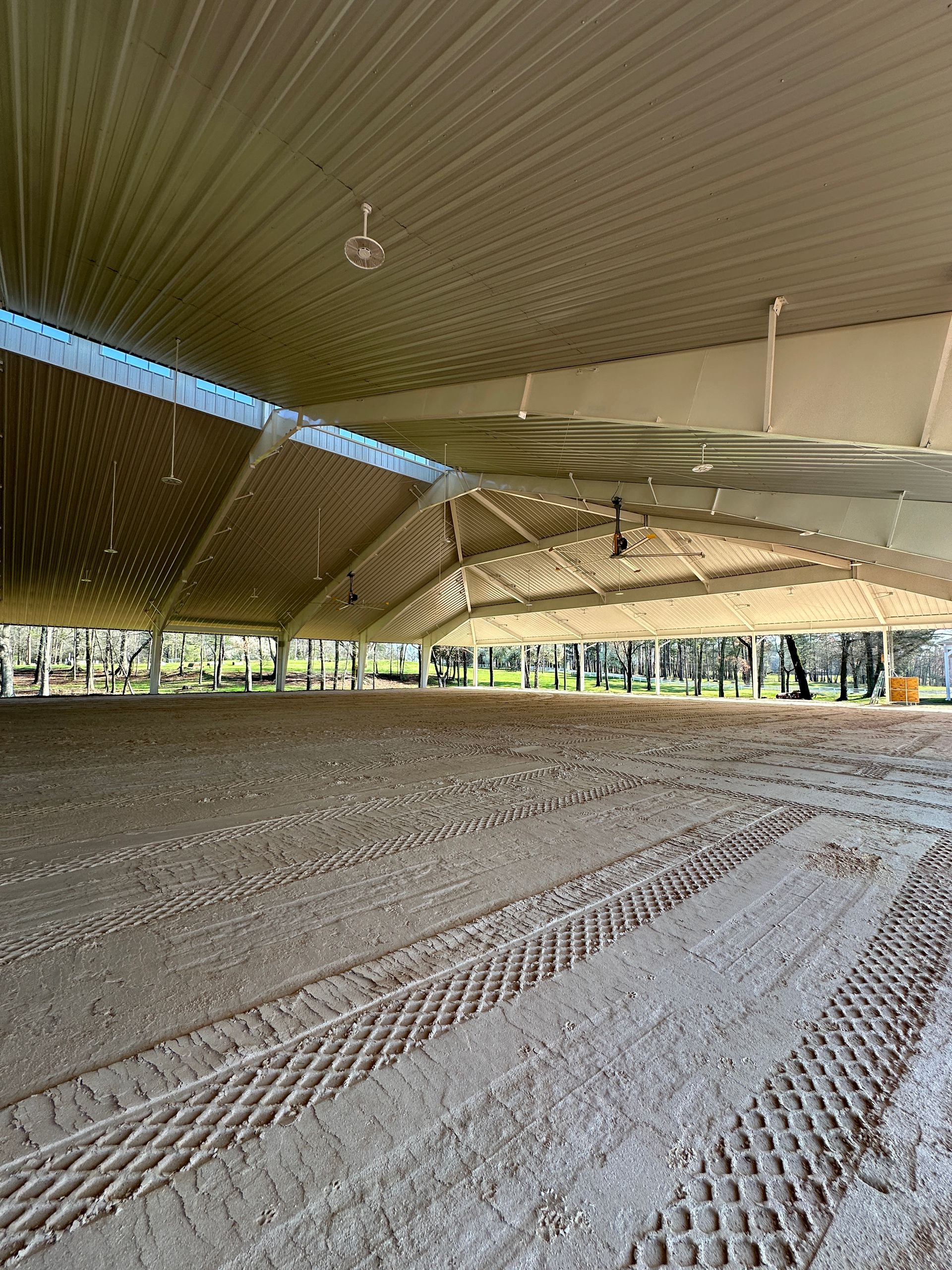 An open-sided indoor riding arena with a textured dirt floor, tire tracks, and a high metal roof.