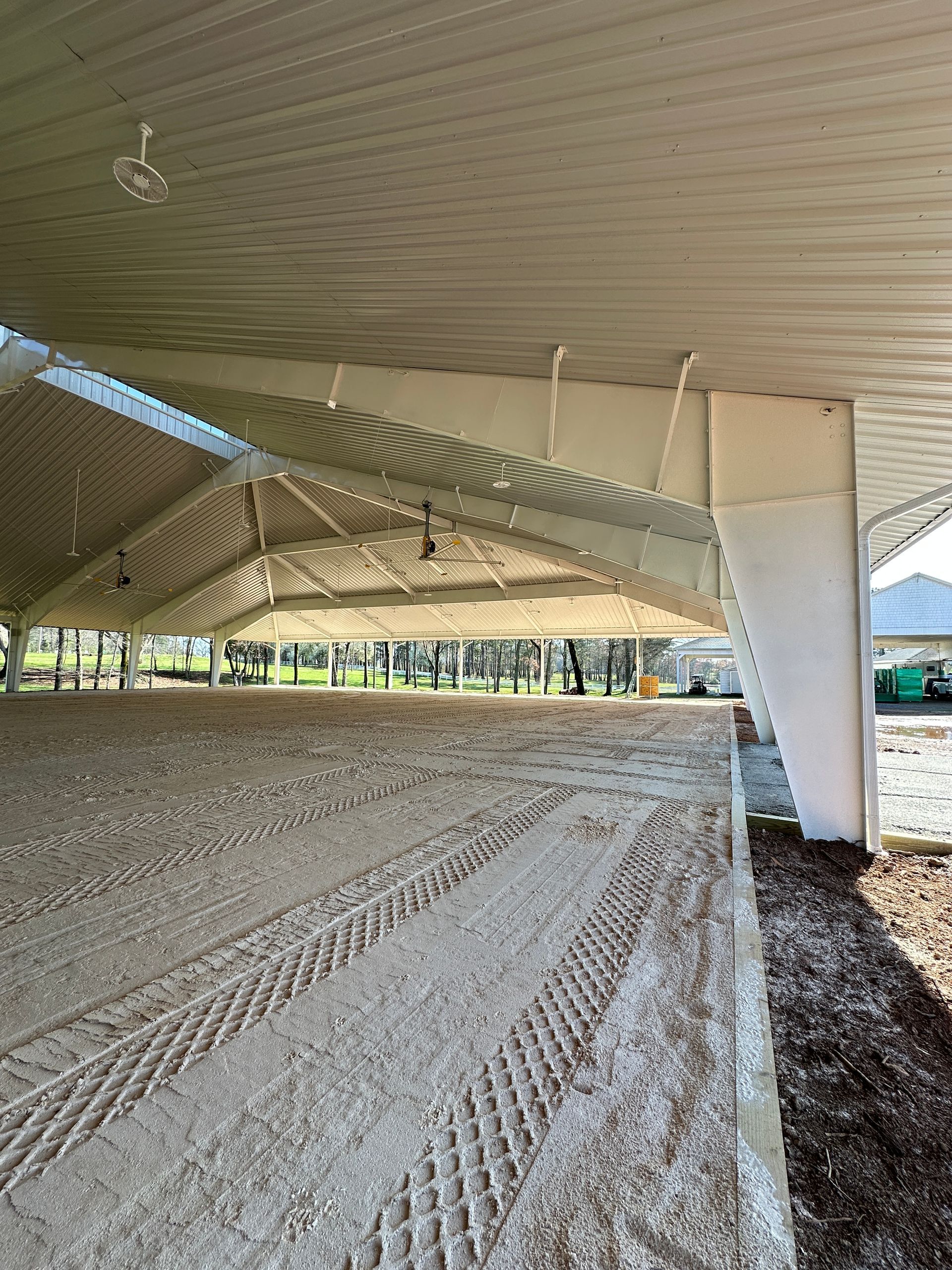 Wide-angle view of a covered riding arena with a large, sand-covered floor featuring tire tracks under a high roof.