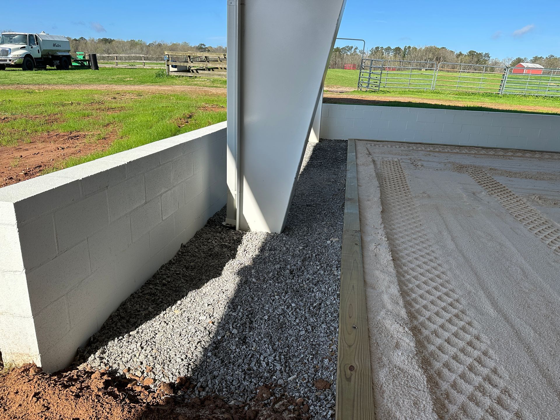 A gravel drainage strip separates a concrete retaining wall from a sandy riding arena under a building overhang.