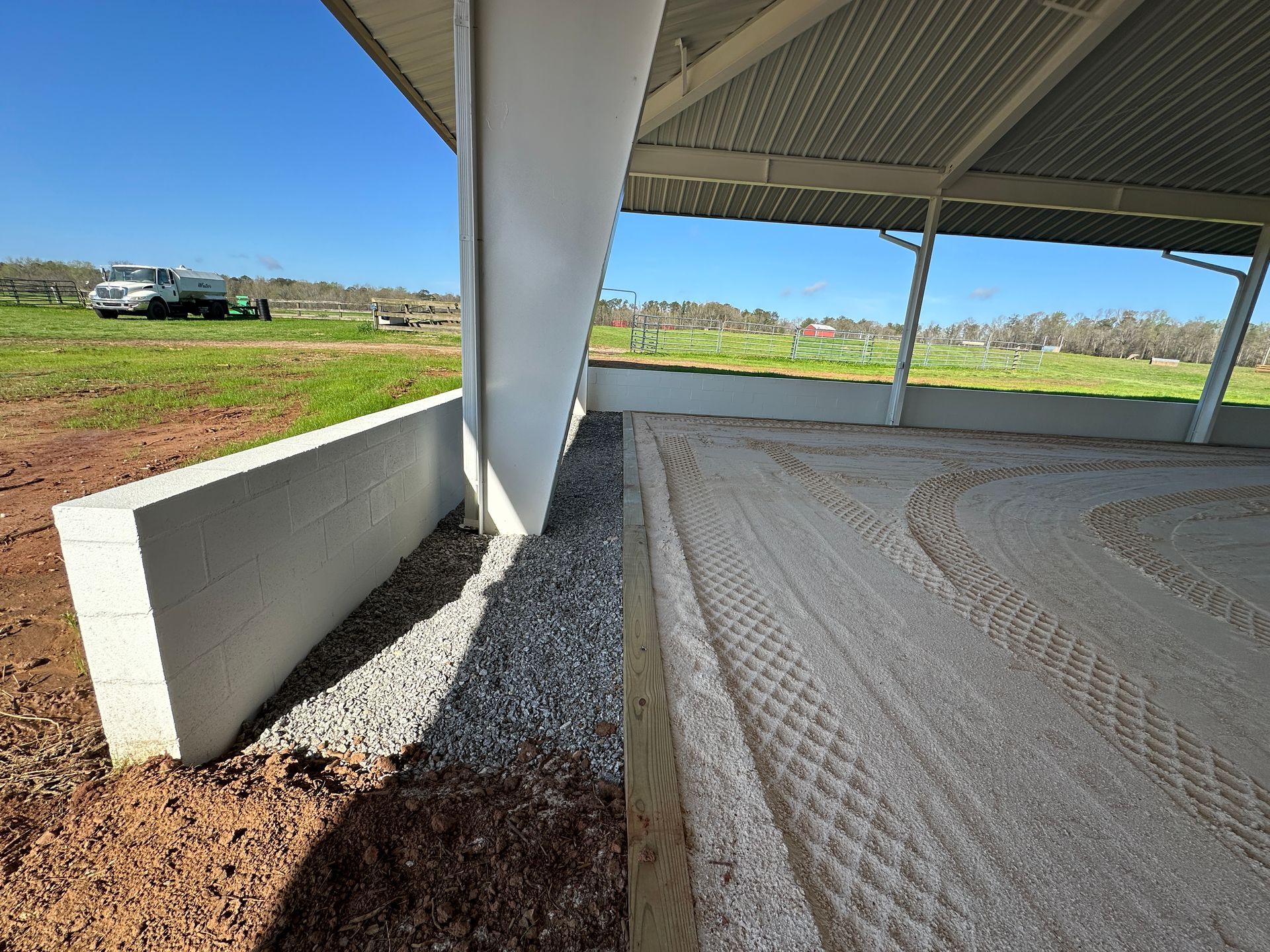 A covered equestrian arena with a sand surface, gravel perimeter, and white retaining walls under a blue sky.