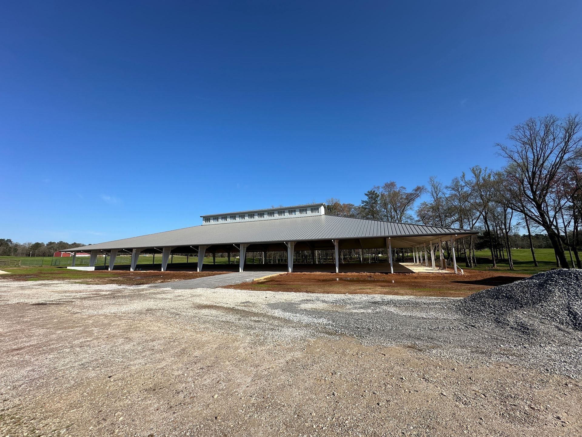 A large, open-air pavilion with a grey metal roof and white support pillars set in a gravel lot under a clear blue sky.