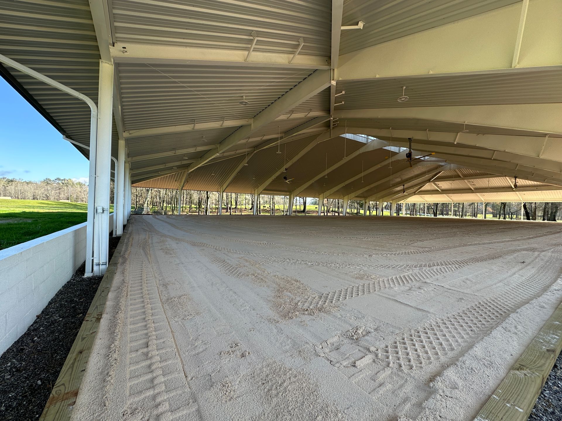 Covered sandy arena with beige roof, white barrier, and open sides letting in daylight.