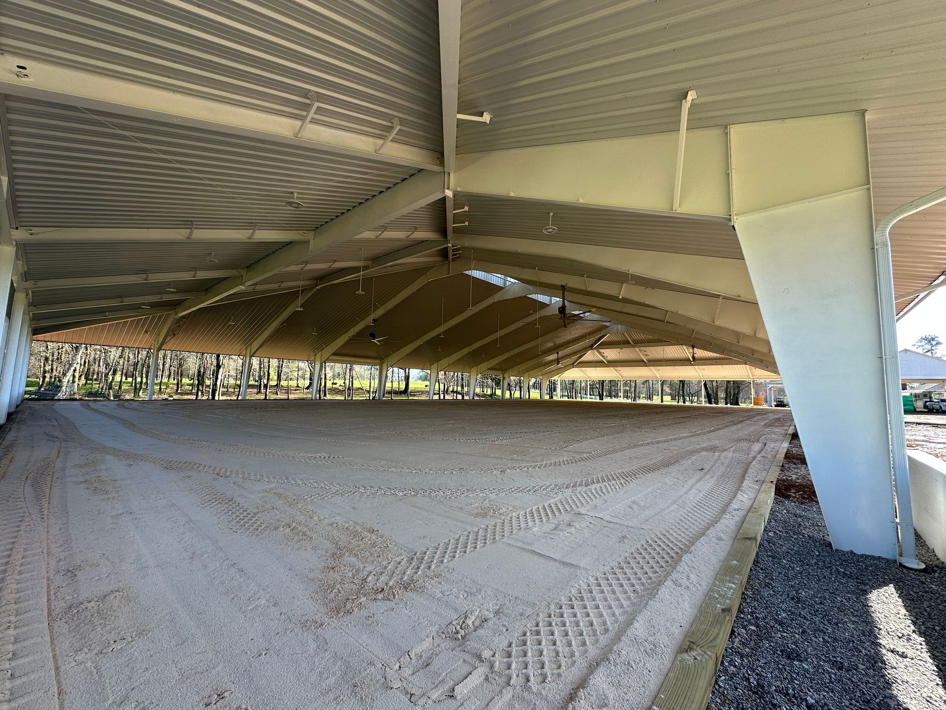 Large, covered riding arena with a sandy surface, open sides, and a white, angular metal roof supported by white pillars.