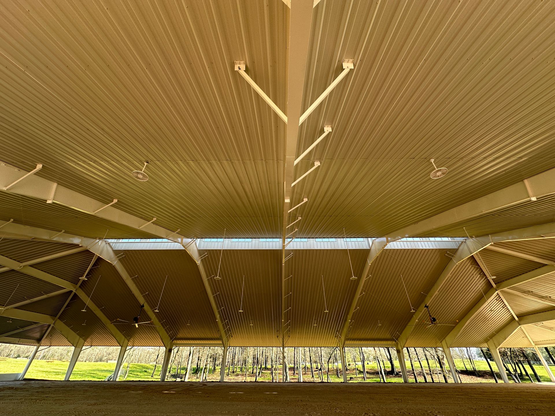 The interior of a large, open-air pavilion with a vaulted metal roof, support beams, and a central skylight strip.