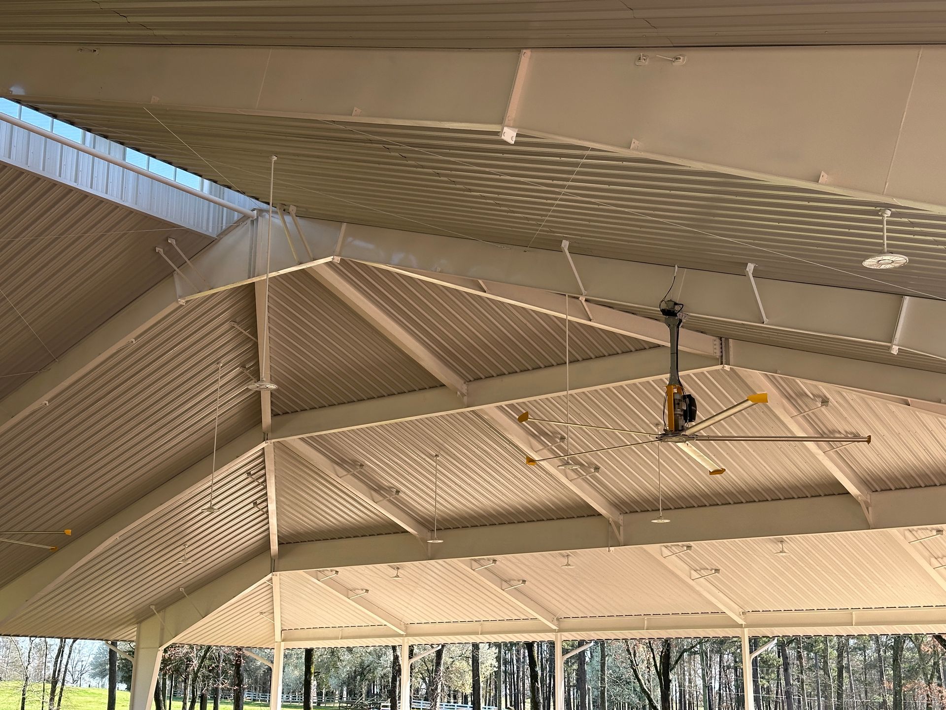 A view of a white, gabled pavilion ceiling with structural beams, a central industrial fan, and mesh netting.
