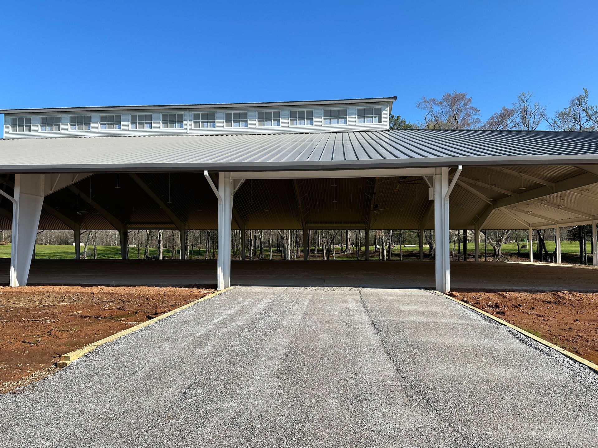 A gravel path leads to an open-air pavilion with a grey metal roof, set against trees and a clear blue sky.