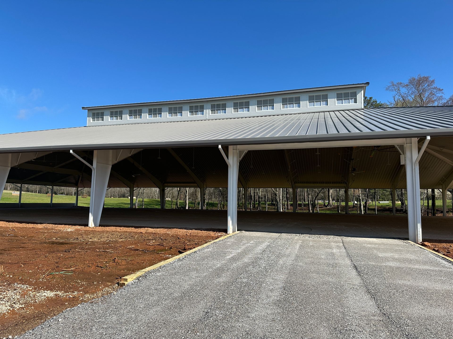 A large, open-air pavilion with a grey metal roof and white support columns, set in a rural landscape under a blue sky.