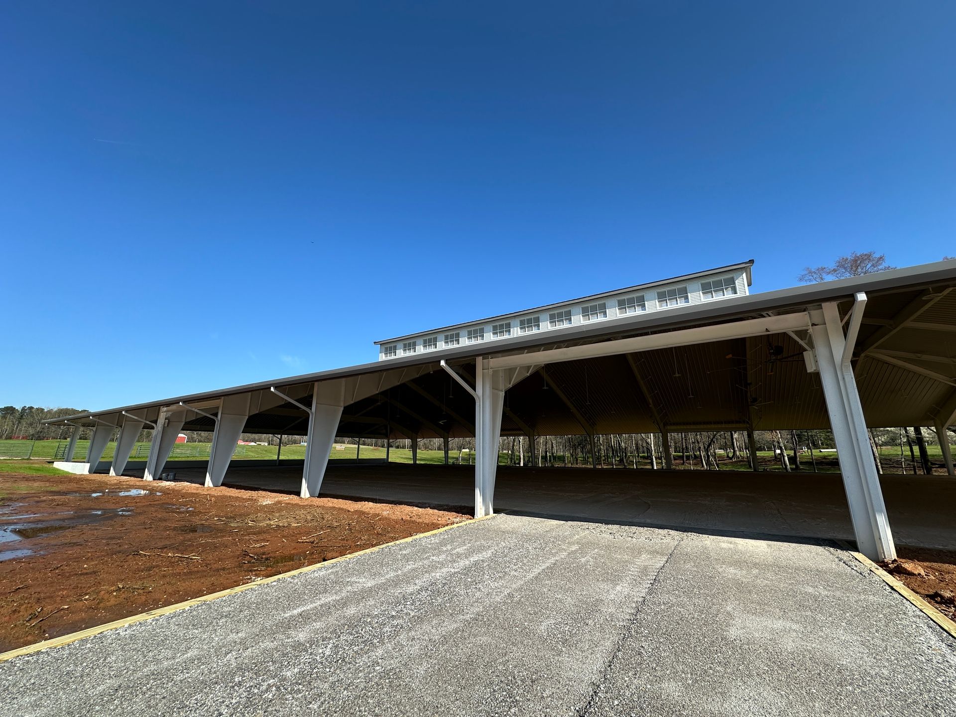 A wide, open-sided pavilion with a metal roof and white support posts, situated on a gravel path next to a field.