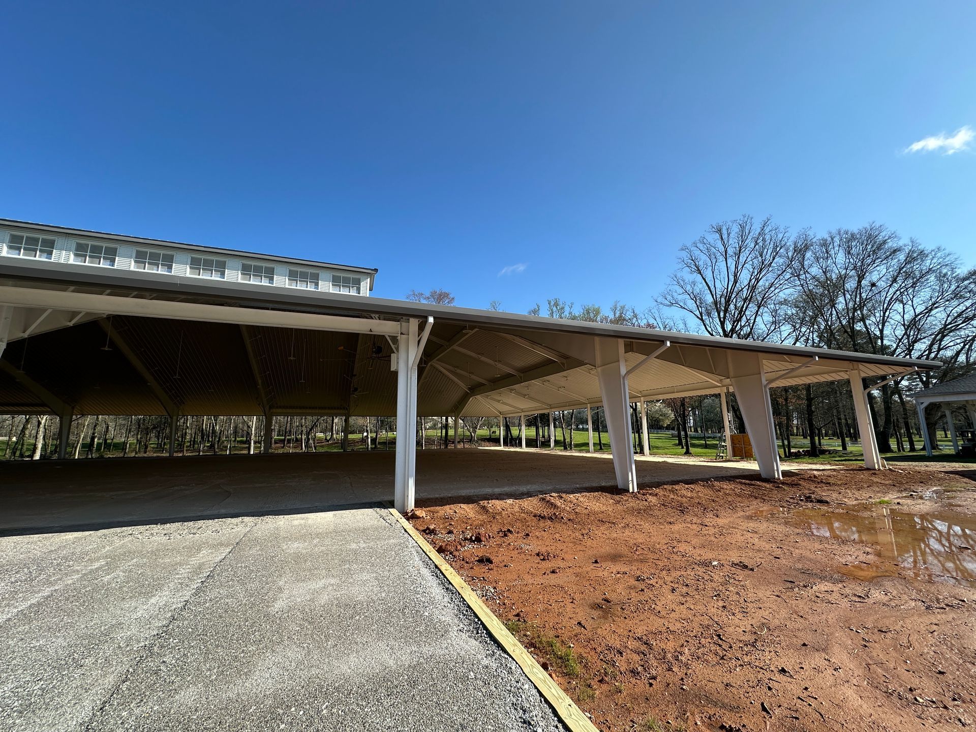 A large, open-air pavilion with white support beams stands over a concrete floor next to a muddy, unfinished ground area.