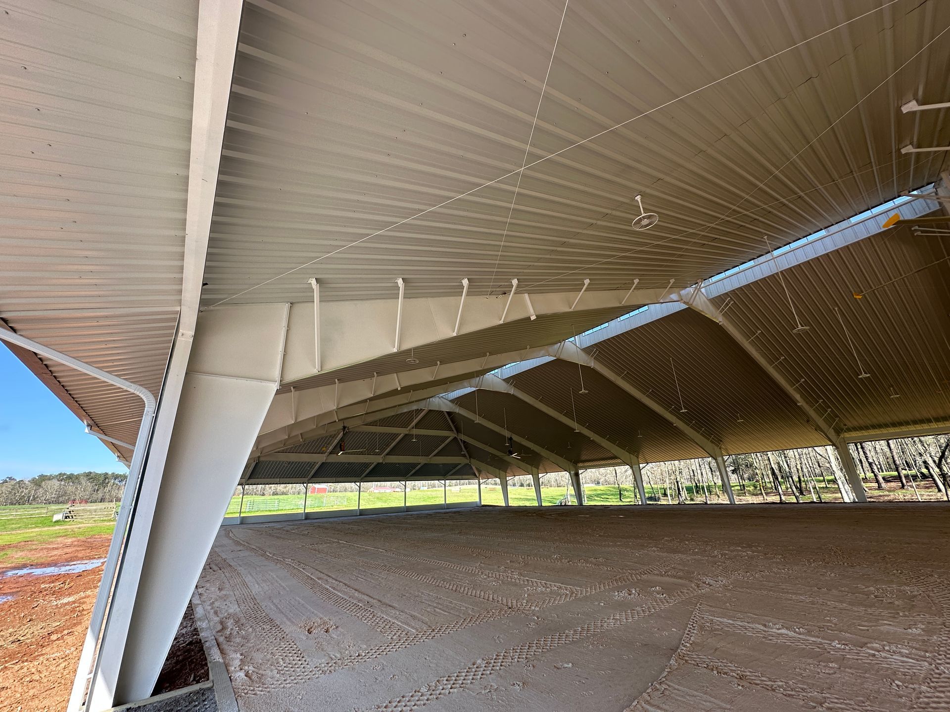 The interior of an open-air riding arena with a metal roof supported by angled white steel columns, viewed from the edge.