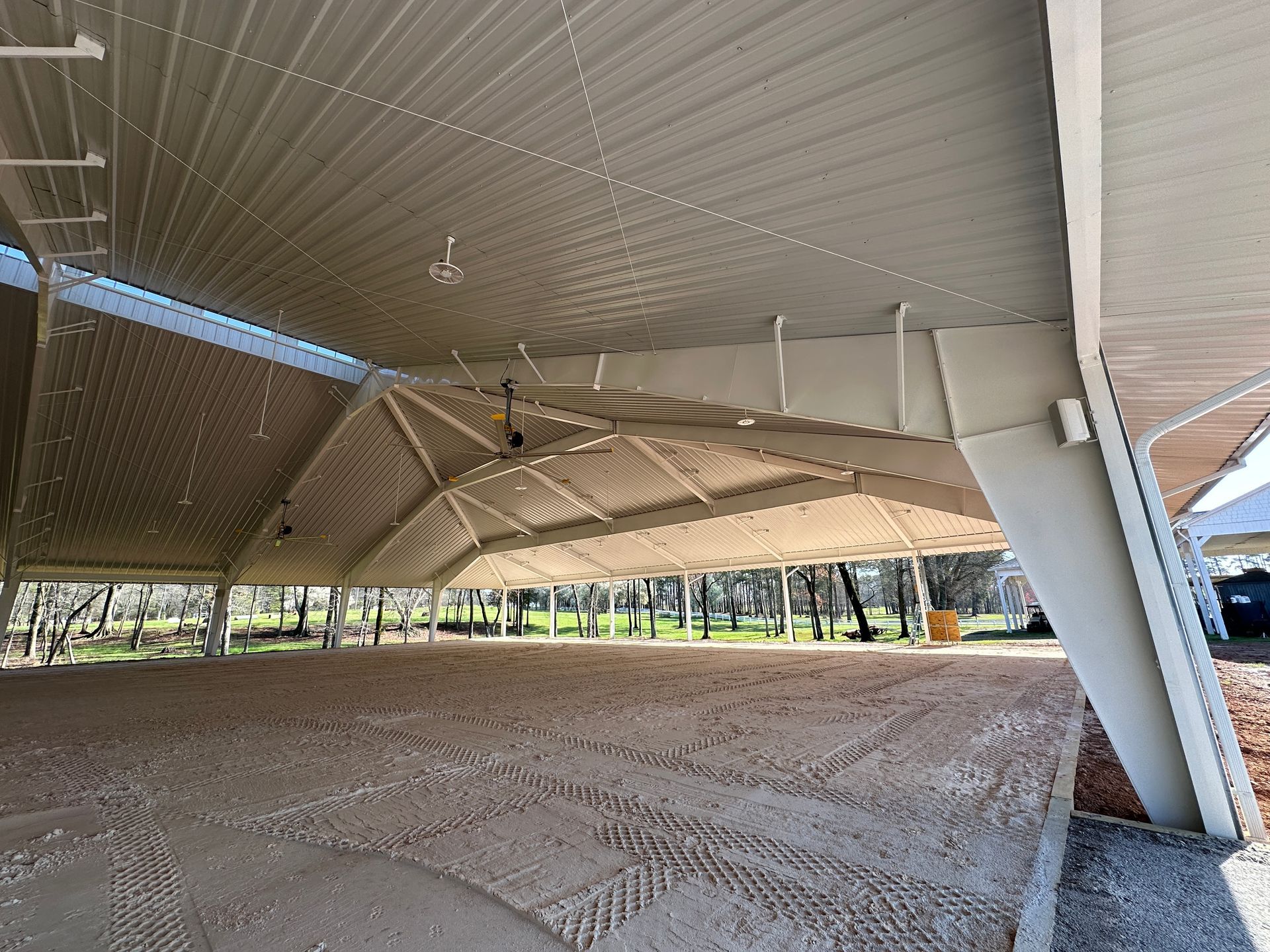 A large, modern open-air pavilion with a metal peaked roof supported by angled concrete pillars over a gravel surface.