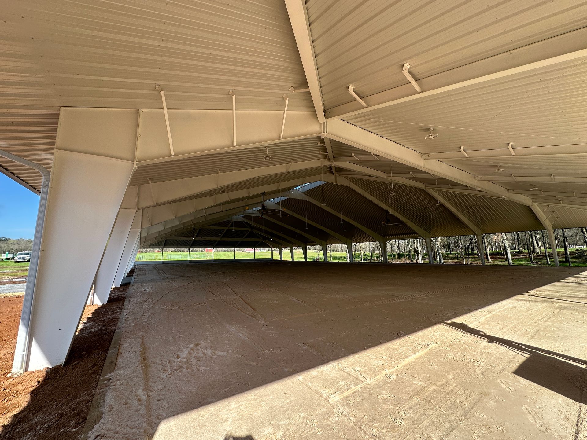 A large, open-air concrete pavilion with a white metal angled roof, supported by white steel beams in a park setting.