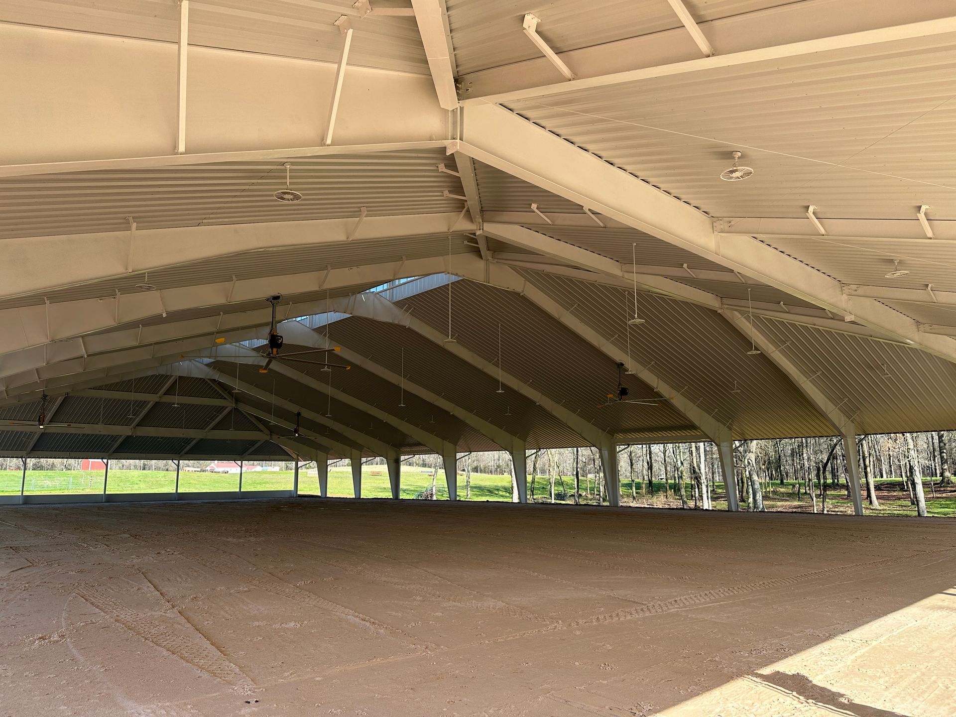 An outdoor, covered horse riding arena with a metal roof, support pillars, and a dirt ground surface.