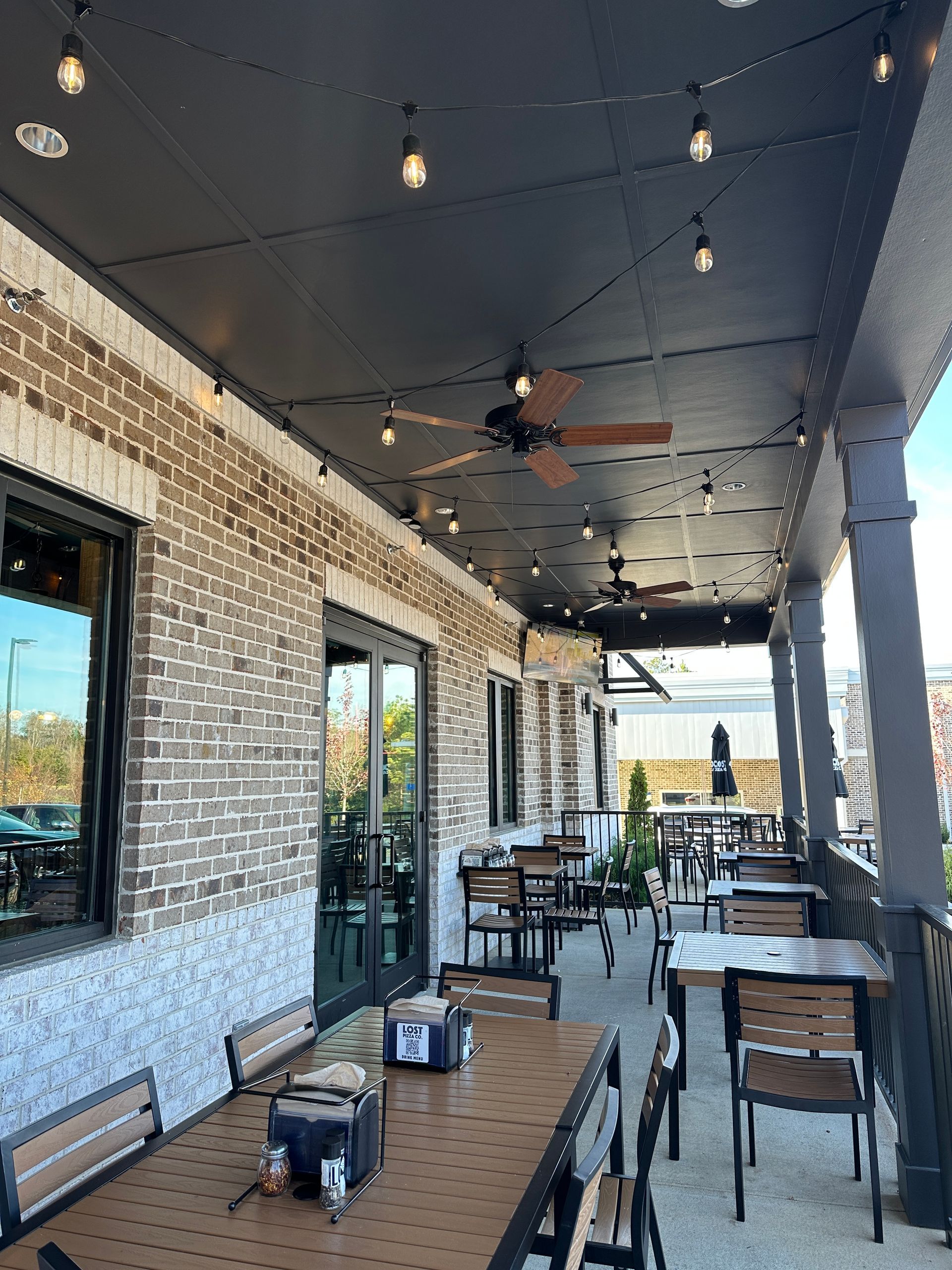 Outdoor restaurant patio with tables, chairs, brick wall, string lights, and ceiling fans.