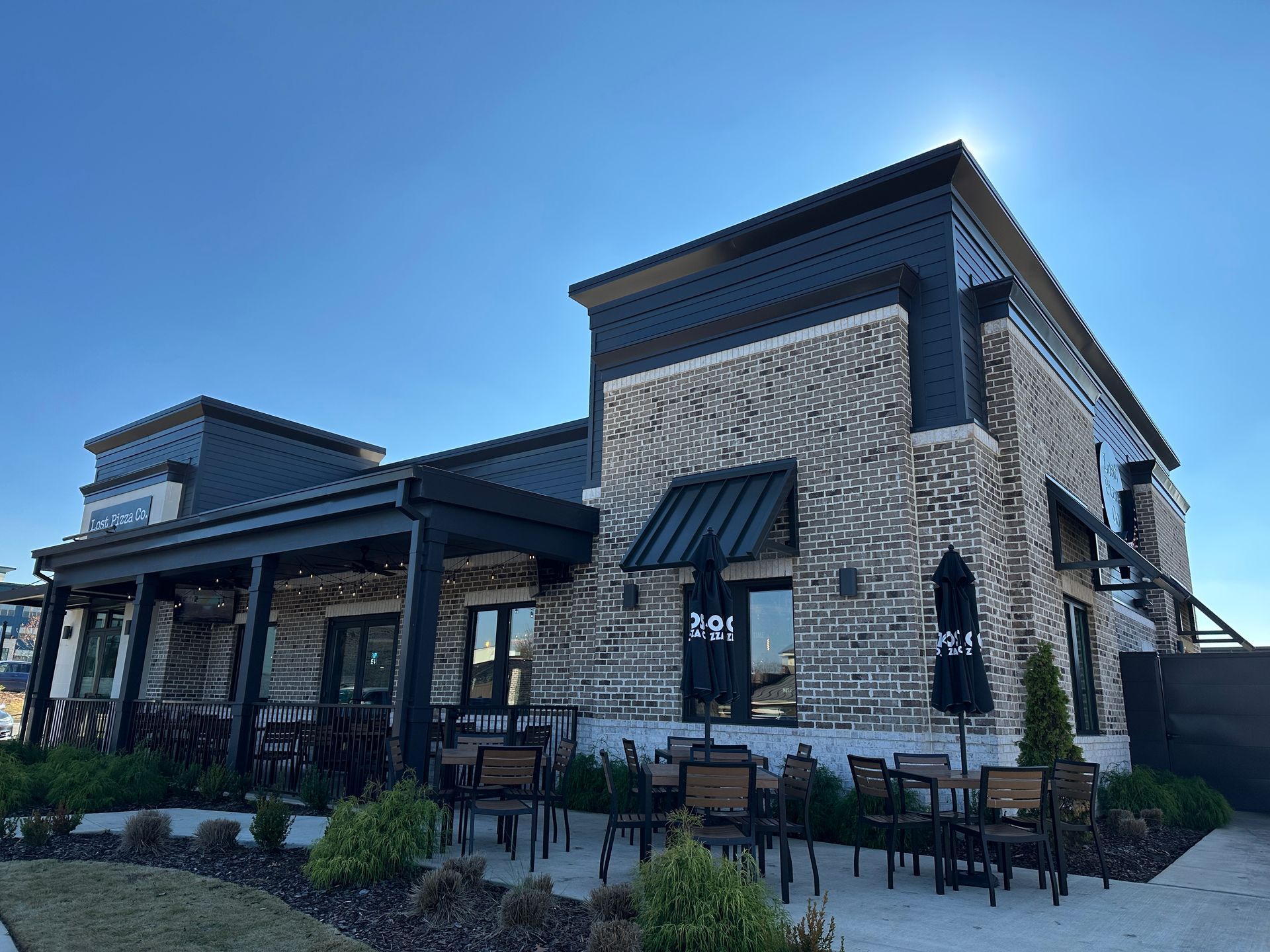 Restaurant exterior with brick and black accents, outdoor seating on patio under awning, sunny day.