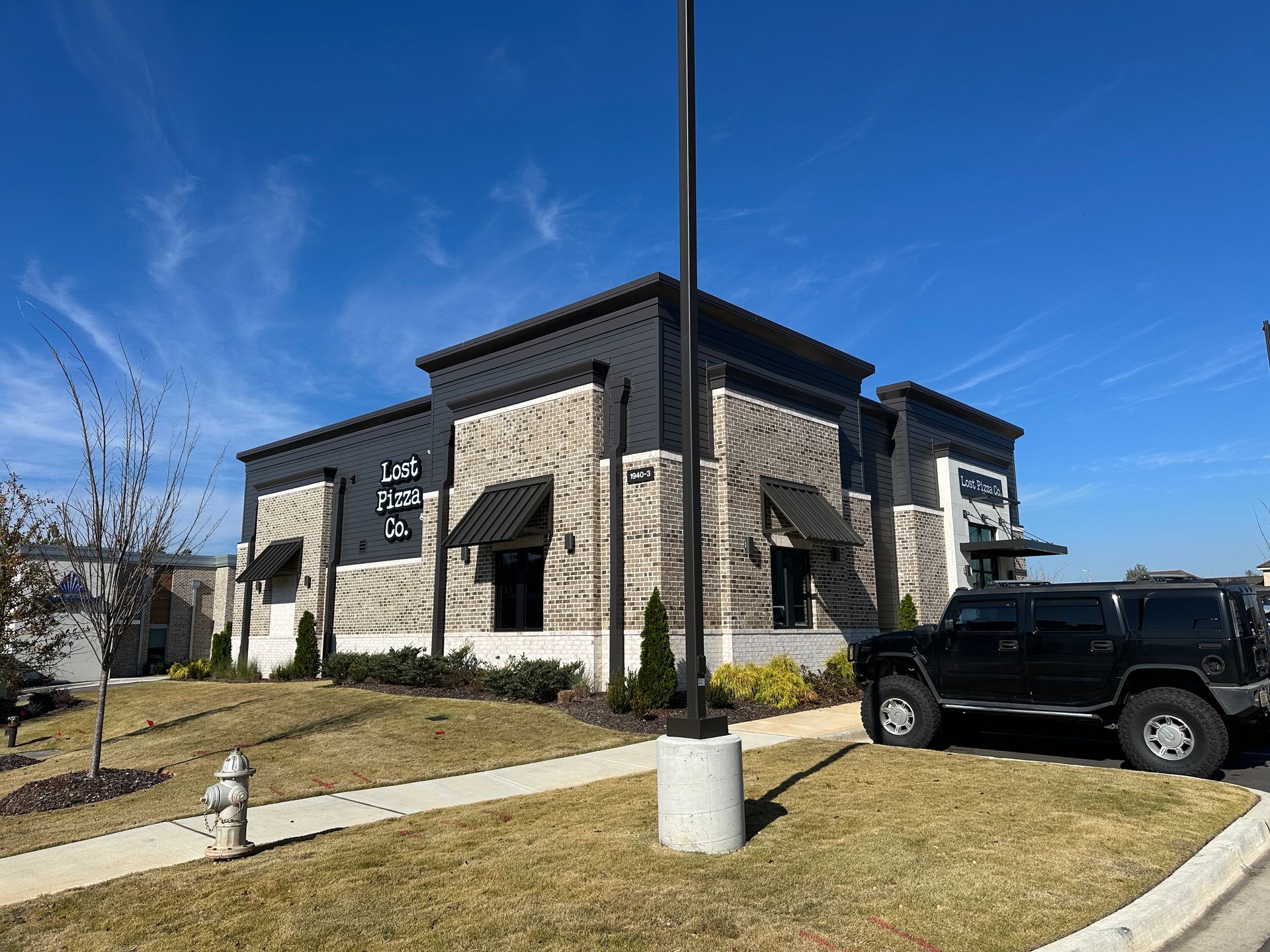 Building with black and tan brick facade, black awnings, and a black SUV parked outside on a sunny day.