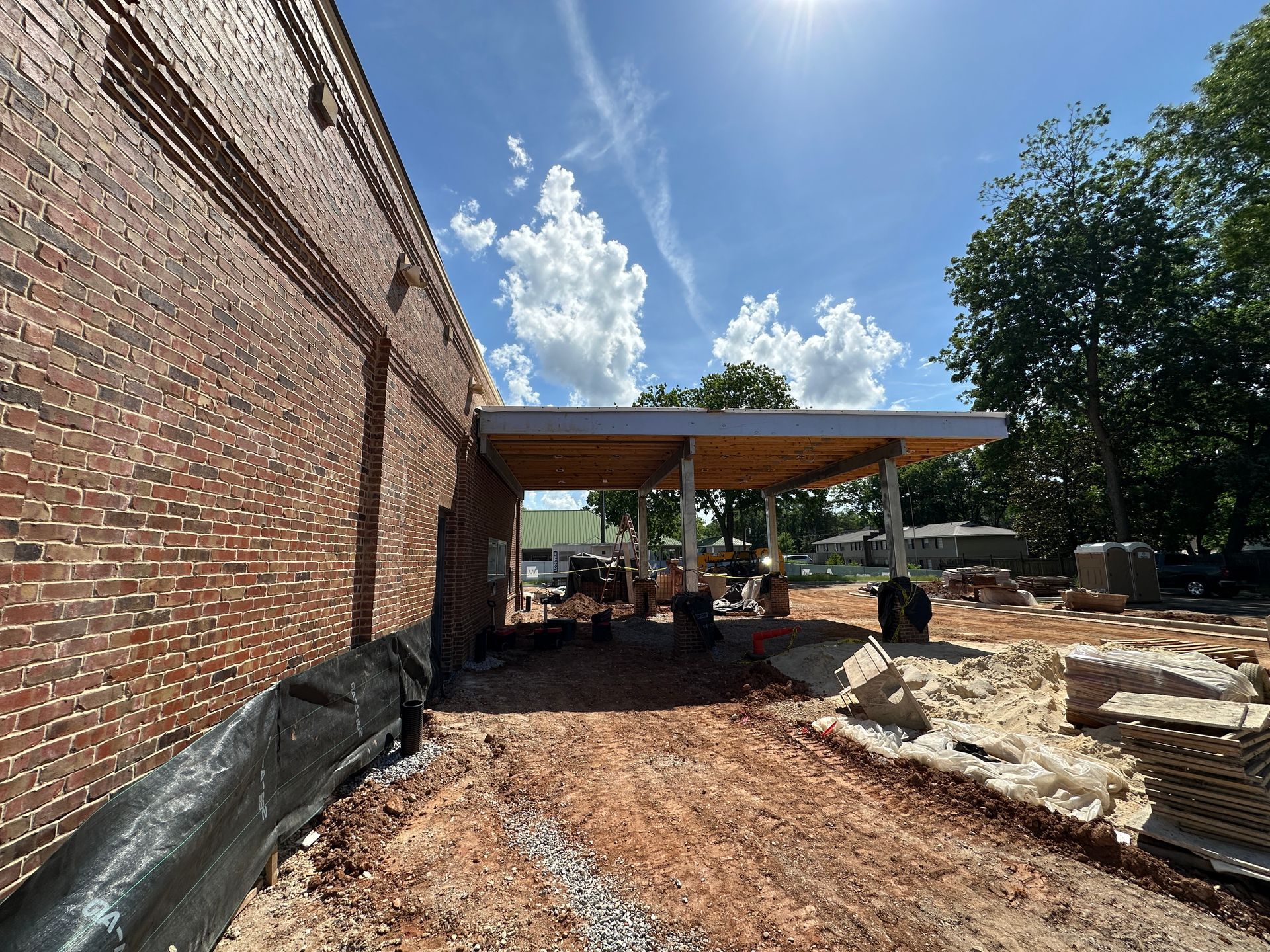 Brick building exterior with covered patio under construction, dirt ground, and scattered materials under blue sky
