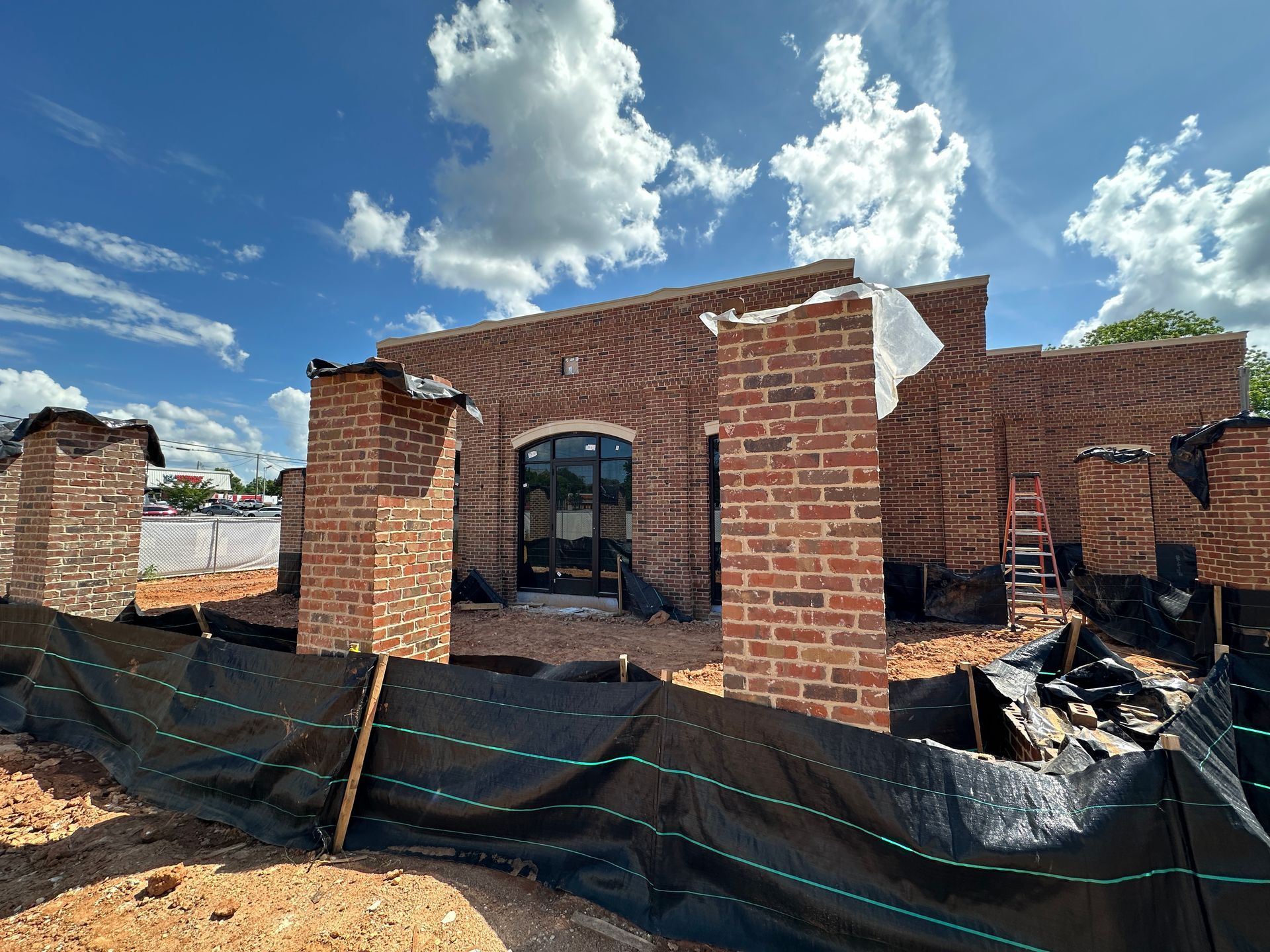Partially built brick house under a blue sky with clouds, surrounded by construction materials and tarp fencing