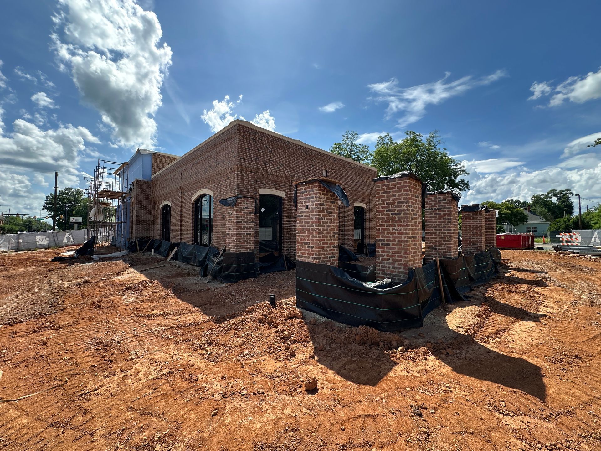 Partially built brick bank with black foundation wrap on a dirt lot under a blue sky