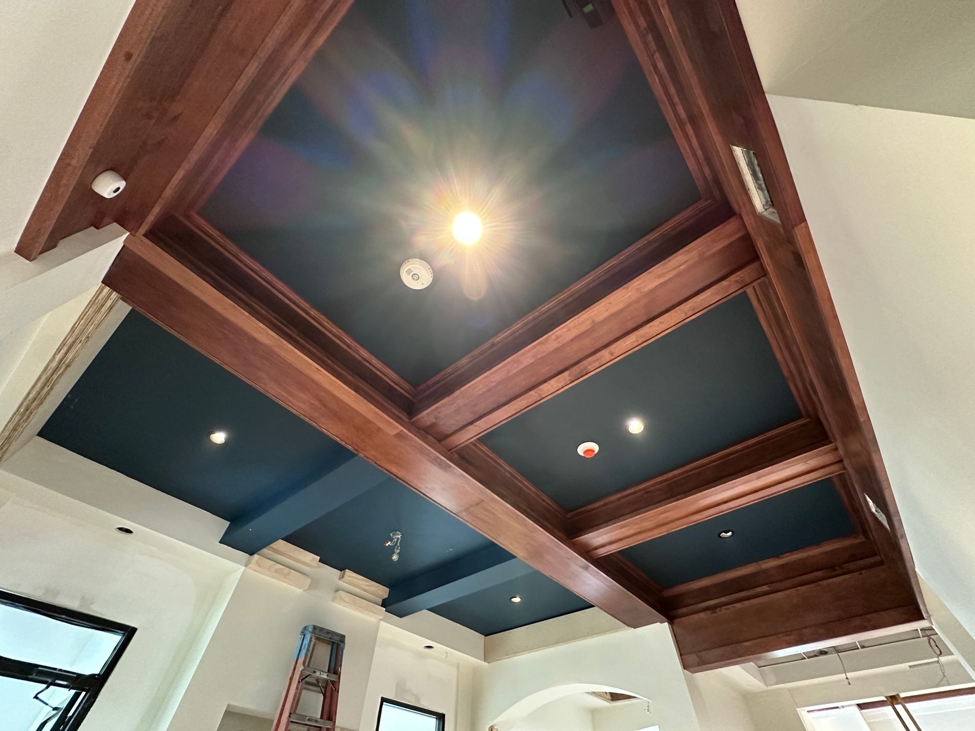 Ceiling with dark blue recessed panels and wood beams, lit by a central bulb in a room under renovation