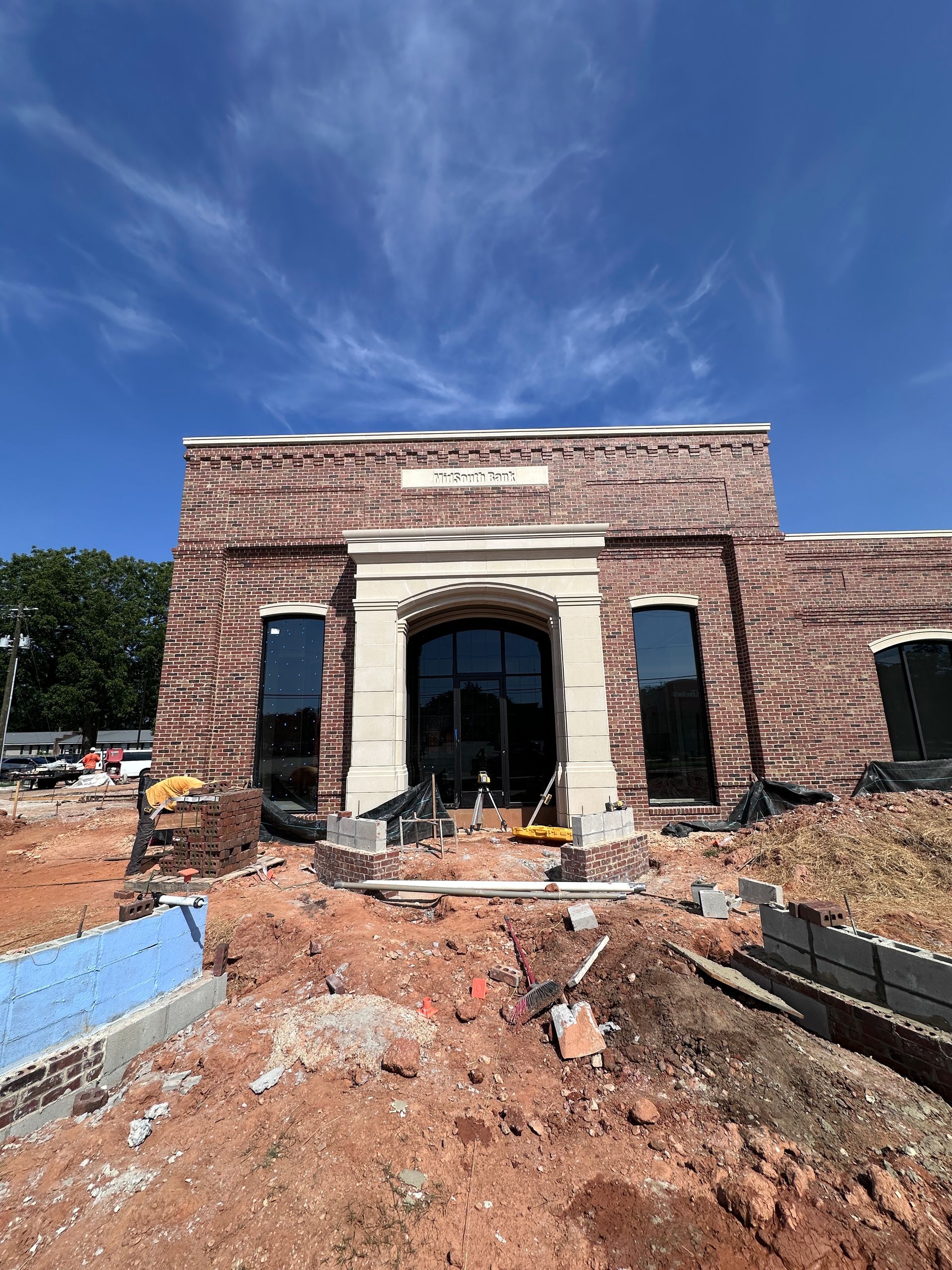 Brick building under construction with large arched entrance, unfinished landscaping, and blue sky.