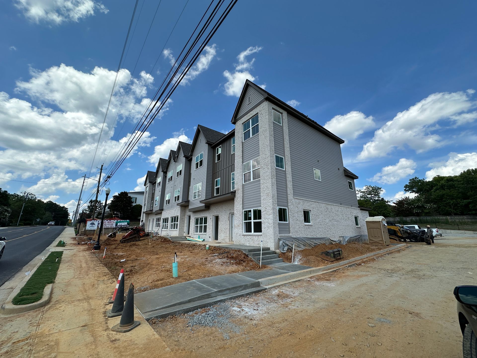 New townhouse row under construction beside a road, with gray siding and orange dirt around the site