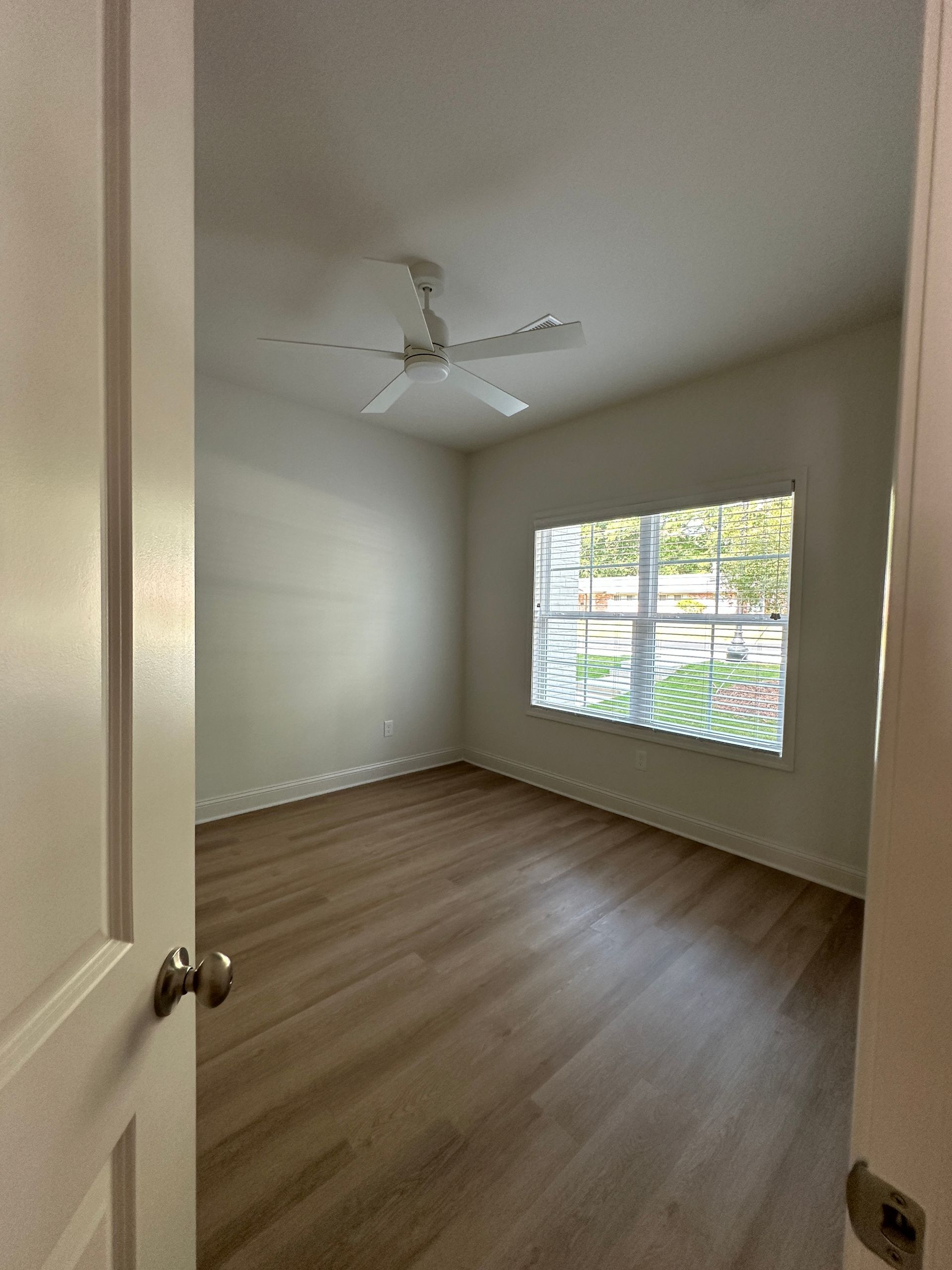 Empty bedroom with white walls, wood floors, ceiling fan, and a window with blinds.