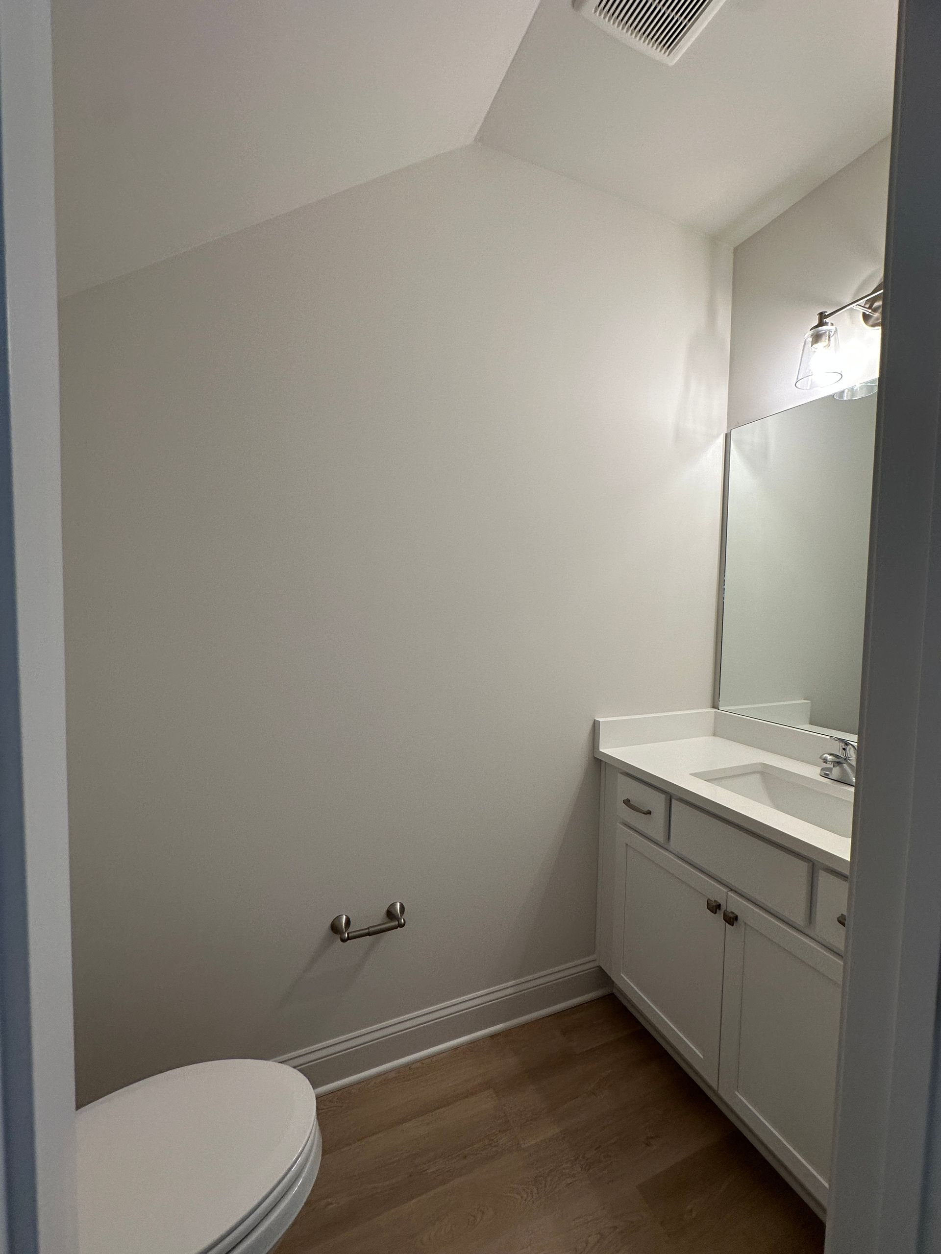 Small white bathroom with toilet, vanity, mirror, and wood-look floor.