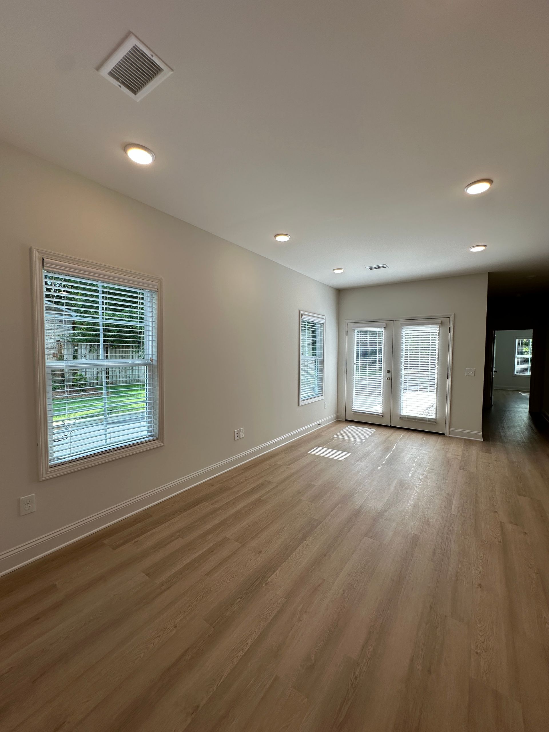 Empty bright living room with hardwood floors, recessed lights, and large windows and glass doors.