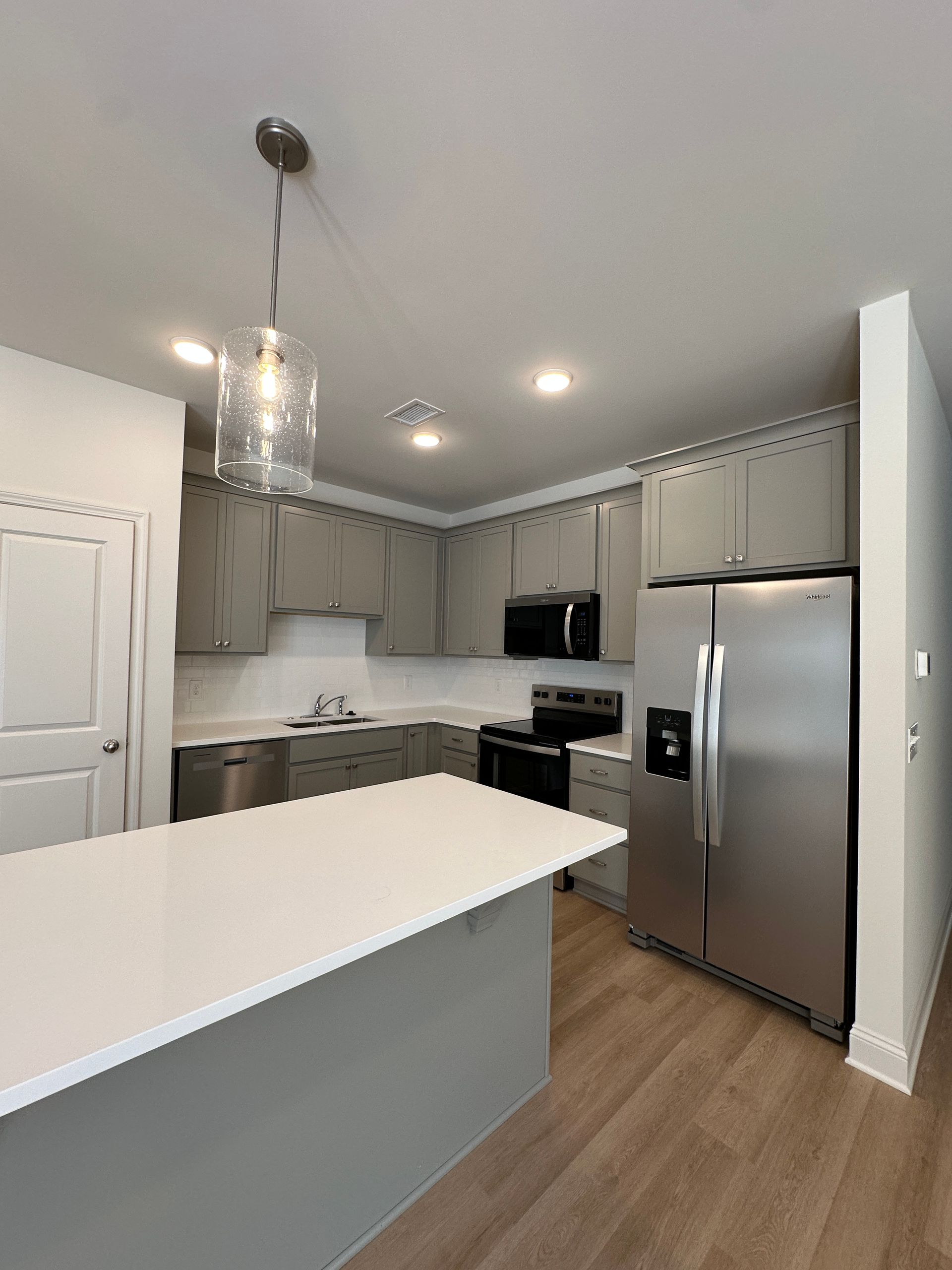 Modern kitchen with white island, gray cabinets, stainless-steel fridge, and light wood flooring.