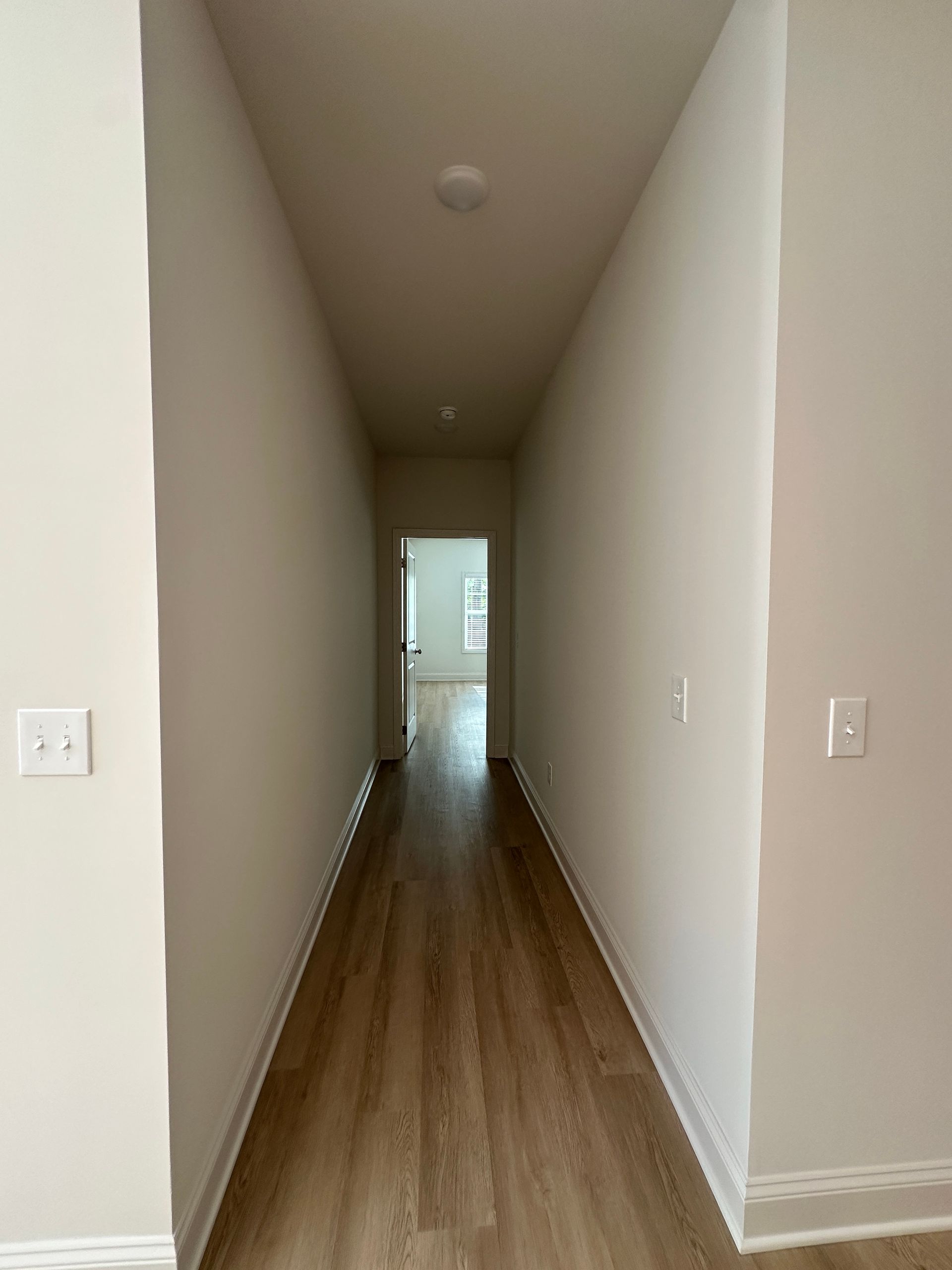 Narrow hallway with beige walls and wood floor, leading to a bright doorway at the end