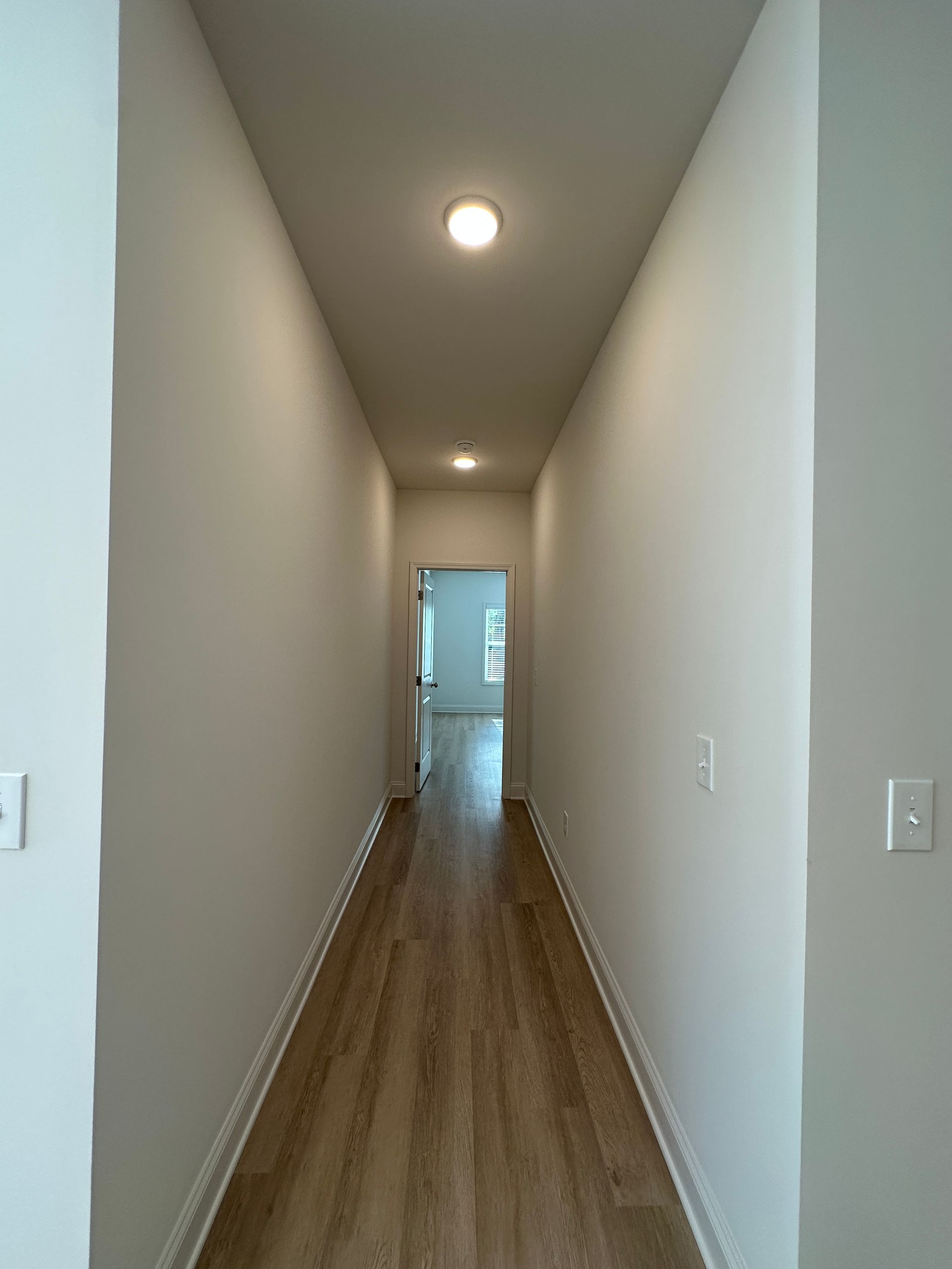 Long, narrow hallway with wood floor, white walls, and ceiling lights, leading to a doorway.