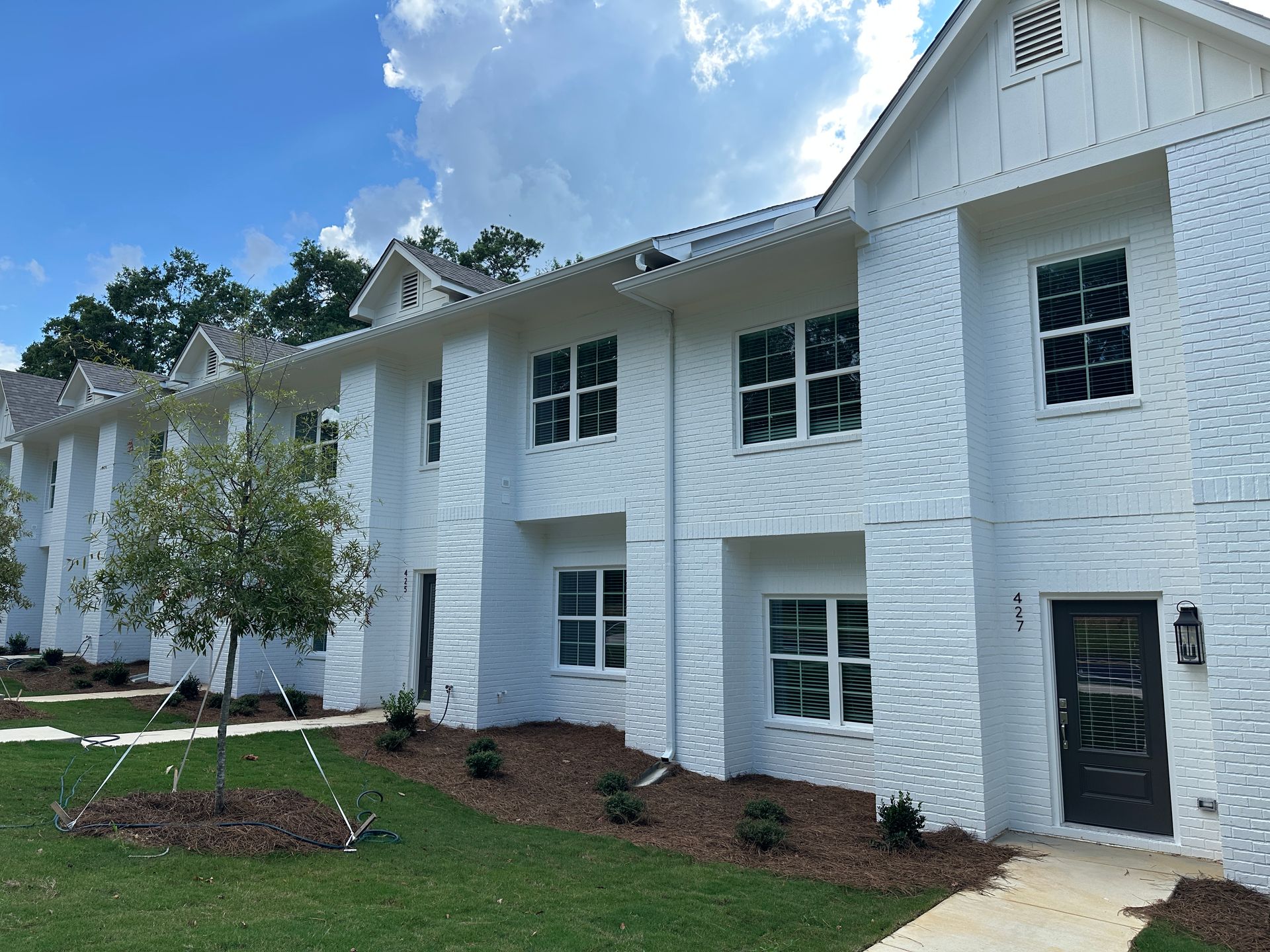 White two-story apartment building with dark shutters, green lawn, and a small tree under a cloudy sky