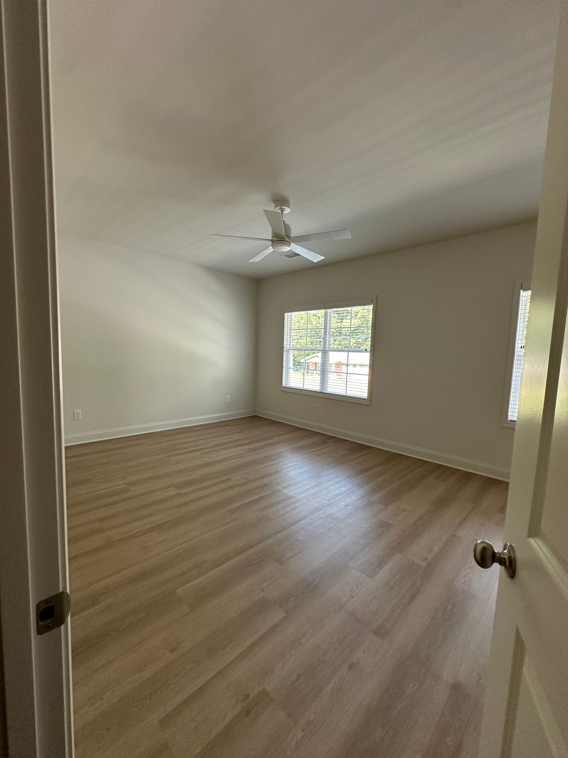 Empty beige bedroom with wood floors, ceiling fan, and a window letting in daylight.