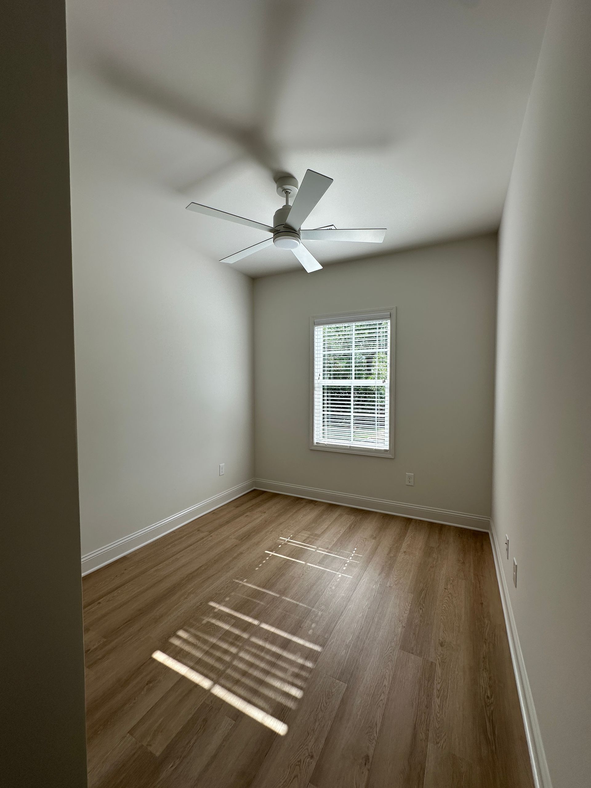 Empty narrow bedroom with white walls, wood floor, ceiling fan, and a window with blinds casting sunlight.