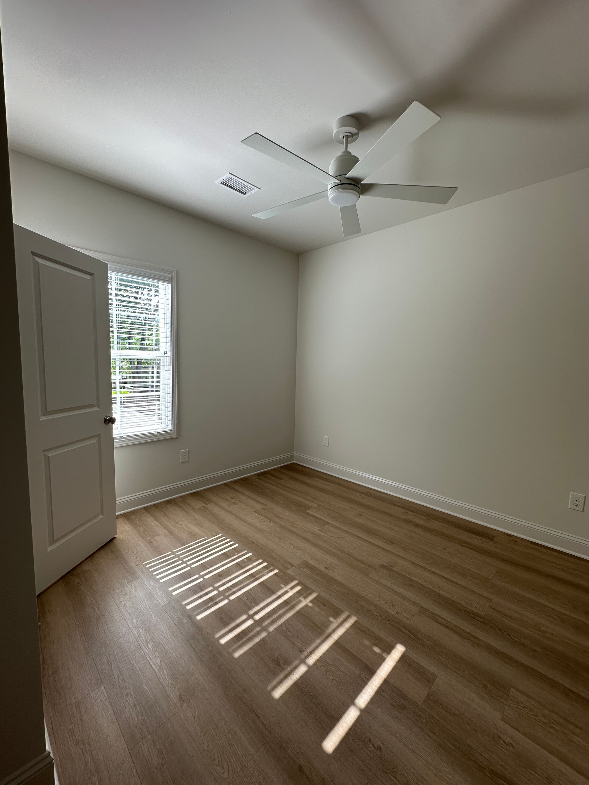 Empty beige bedroom with wood floors, white ceiling fan, and sunlight streaming through a window.