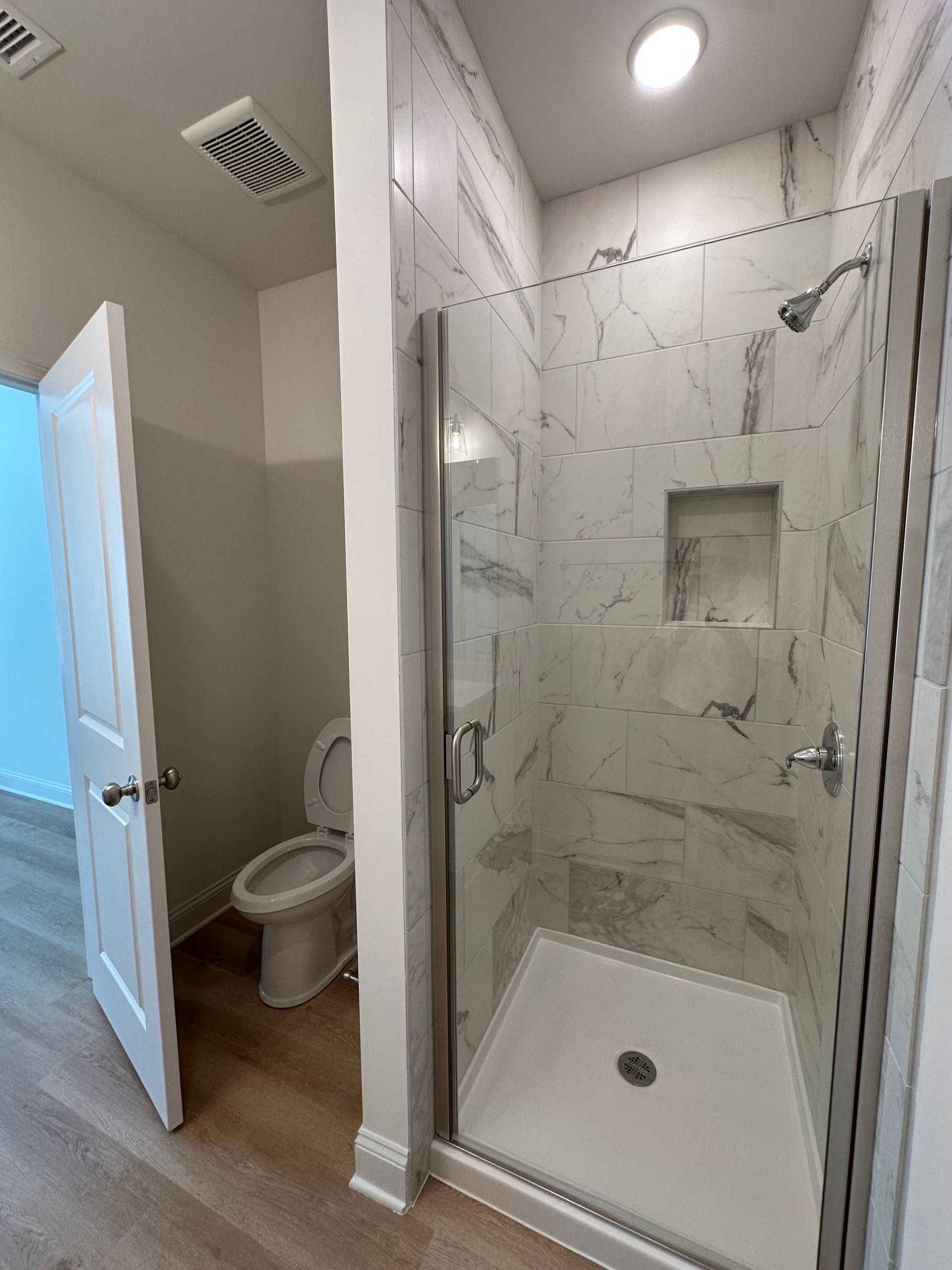 Bathroom with open door and visible toilet beside a glass shower with white tile walls.
