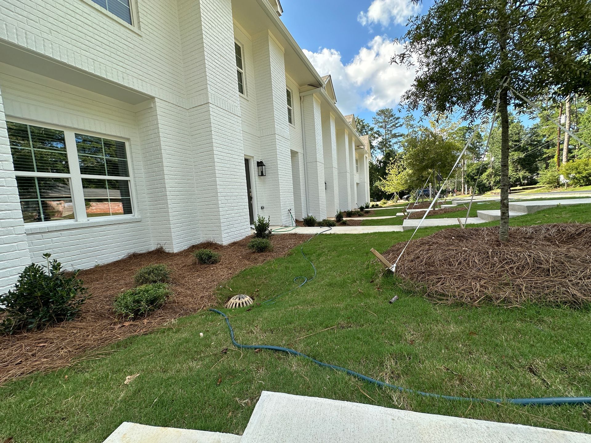 White brick building with windows, grassy lawn, landscaped beds, and trees under a blue sky.