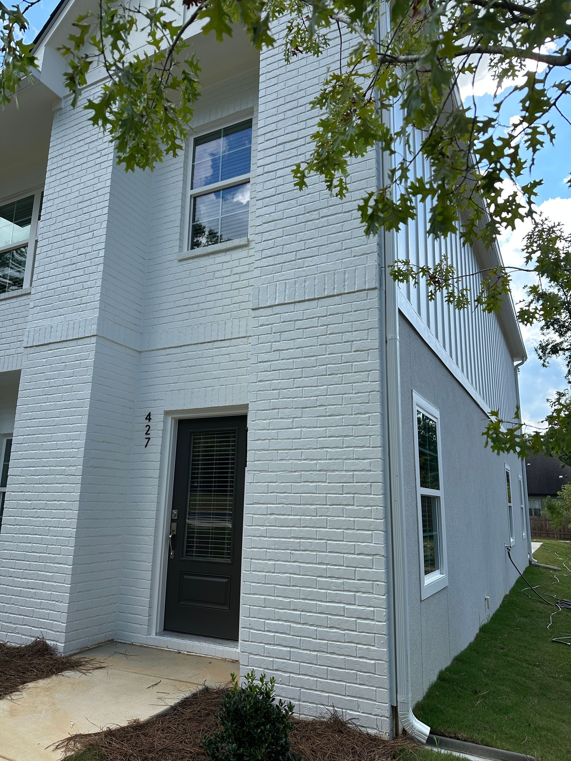 White two-story brick house corner with black door, windows, and tree branches in the foreground.