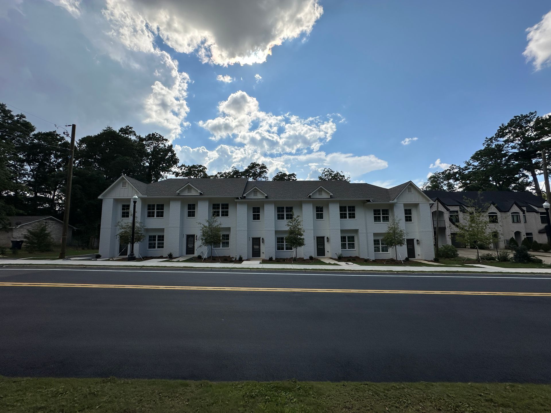 White two-story apartment building with green shutters under a cloudy blue sky, viewed across a road.