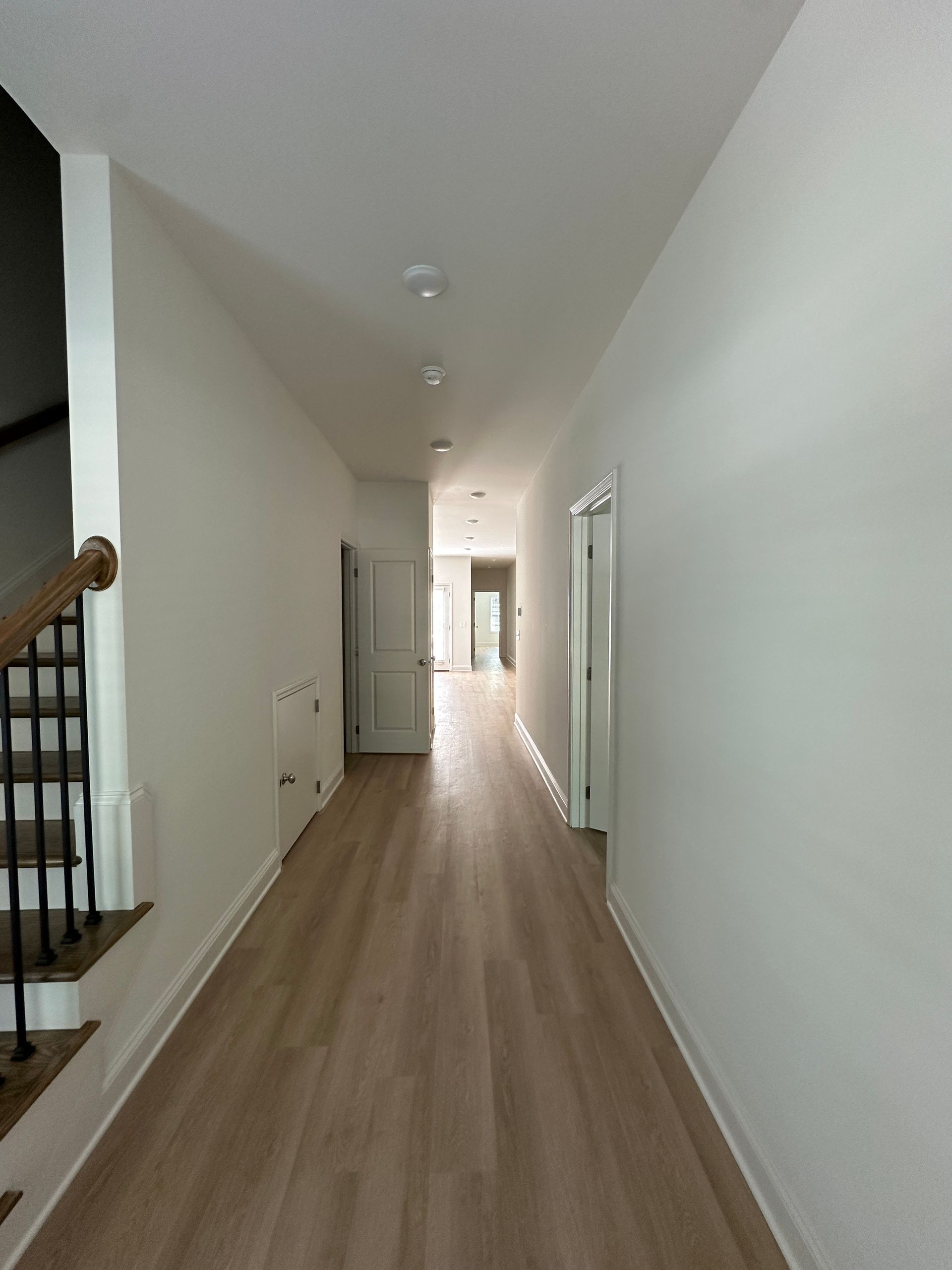 Long narrow hallway with light wood flooring and white walls, seen from one end toward a bright doorway.