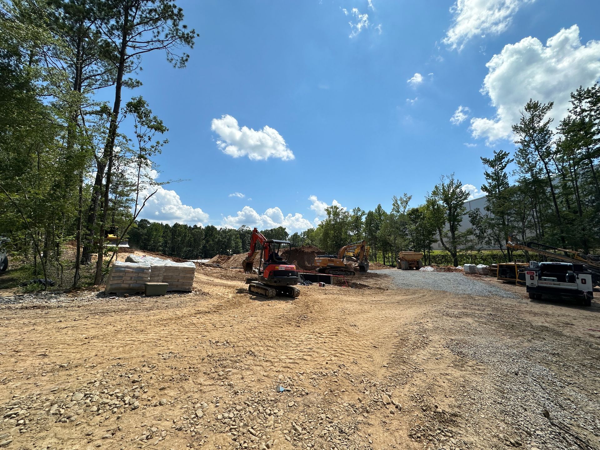Gravel worksite with a red excavator, dirt piles, trees, and a parked truck under a blue sky.
