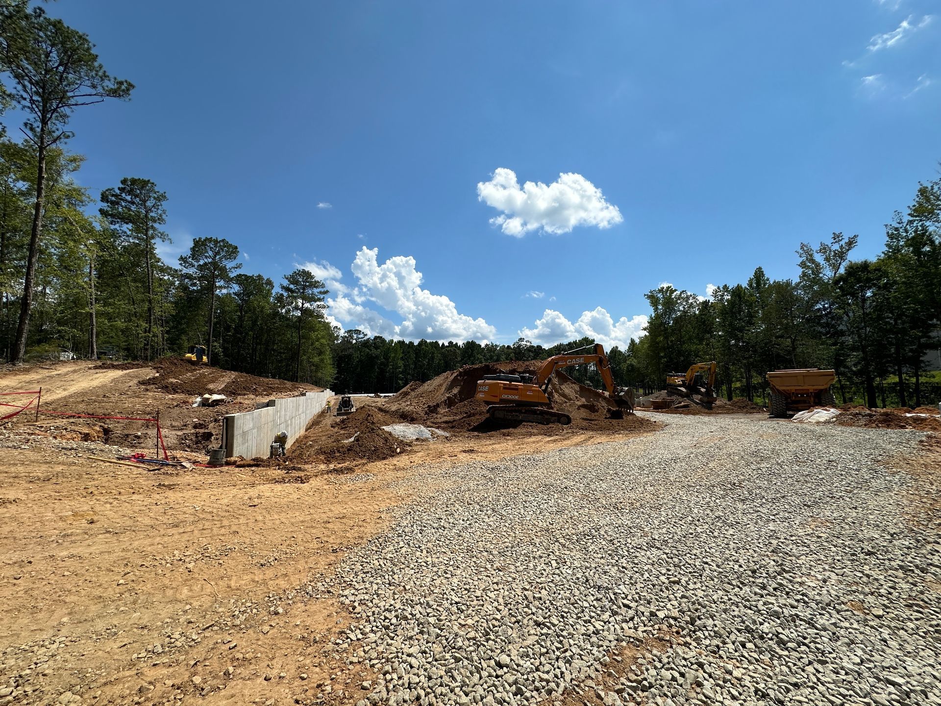 Cleared gravel lot with dirt piles and trees under a bright blue sky with clouds