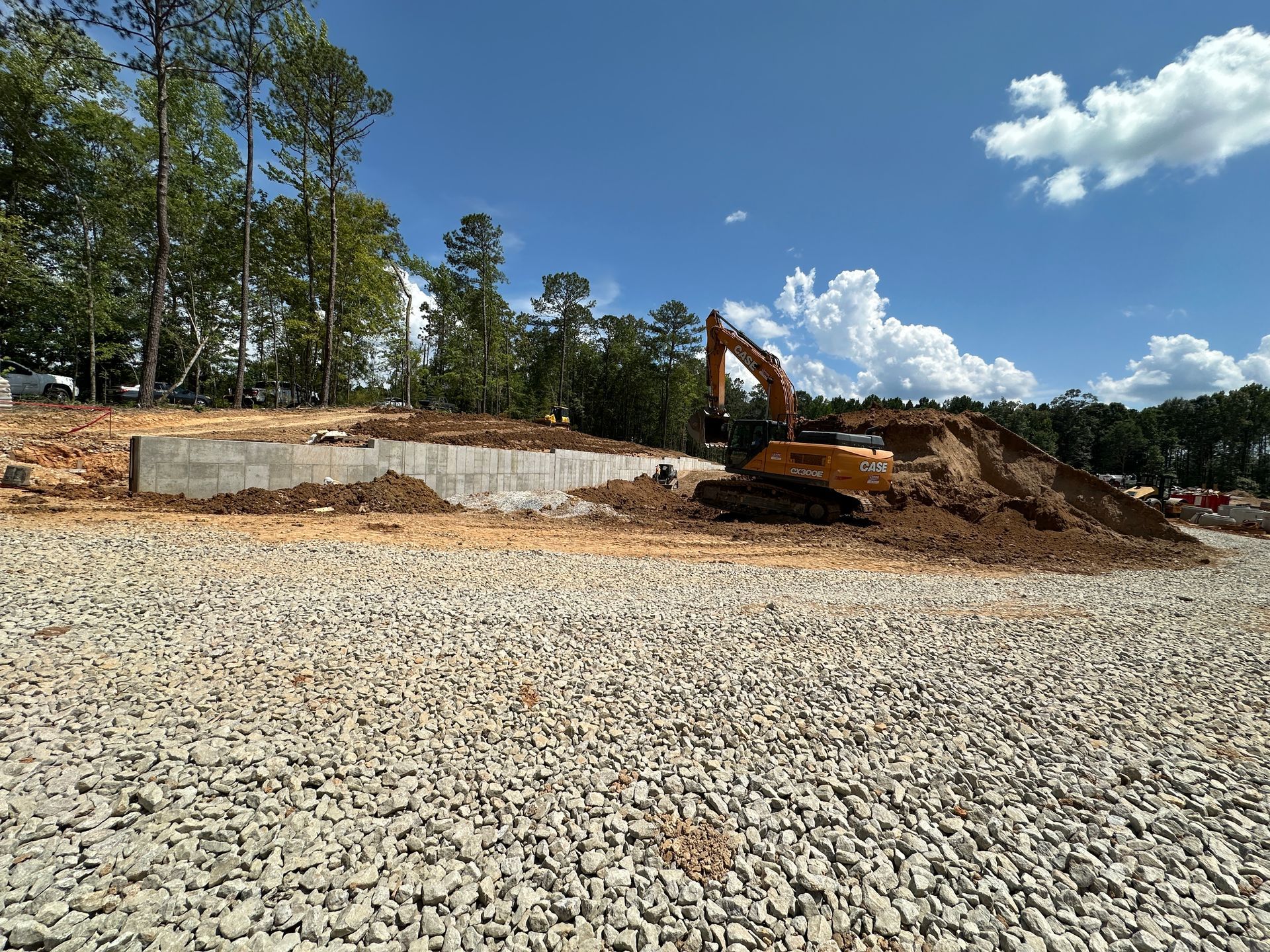 Construction site with dirt piles, heavy equipment, and a tree line under a partly cloudy blue sky