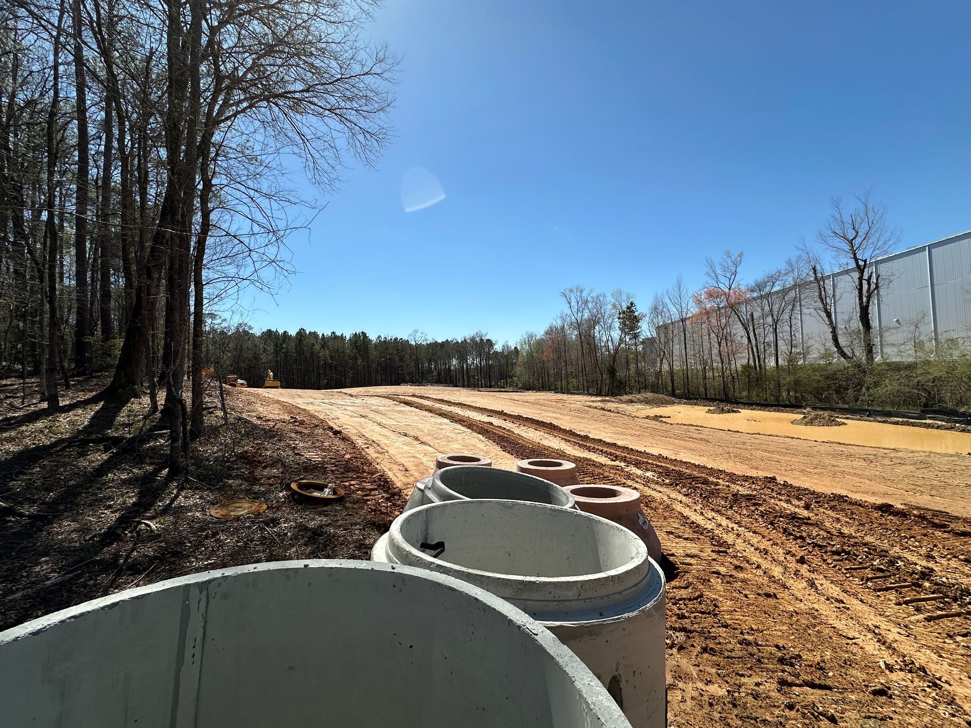 Industrial manufacturing plant under construction with concrete drainage pipes in the foreground and trees along the sides