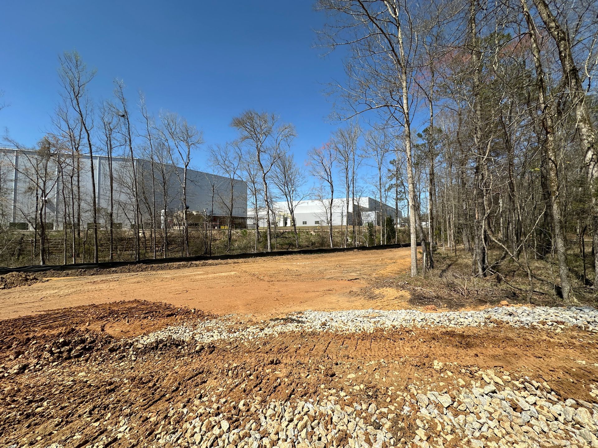 A dirt construction site with a dark silt fence, flanked by bare trees under a clear blue sky, with warehouses in the rear.