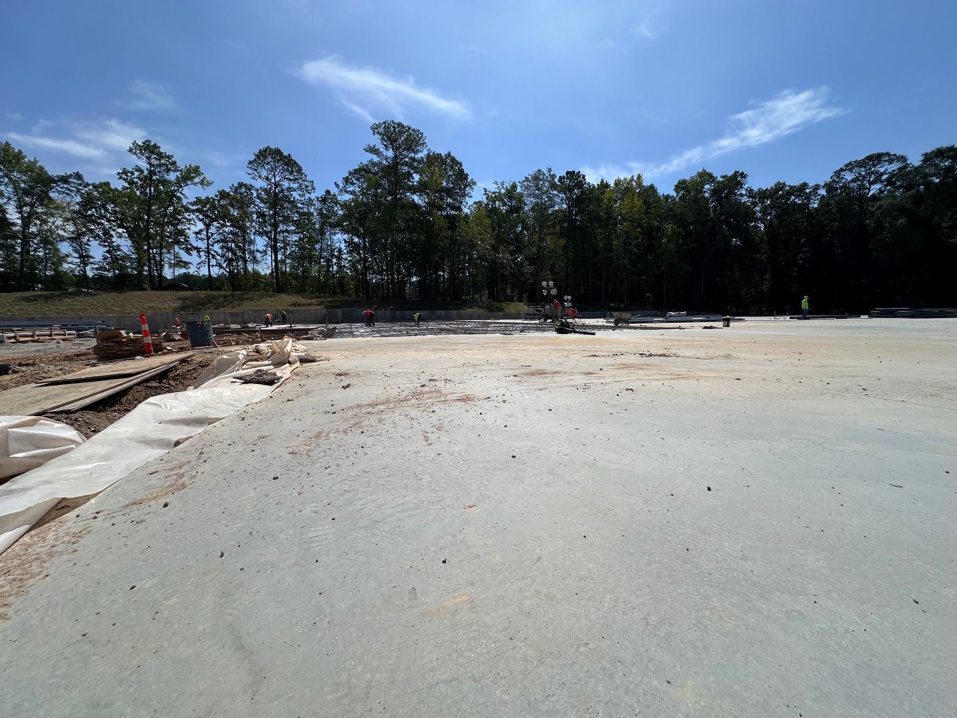 Wide sandy beach with a tree line and blue sky, with a few people and construction materials on the left